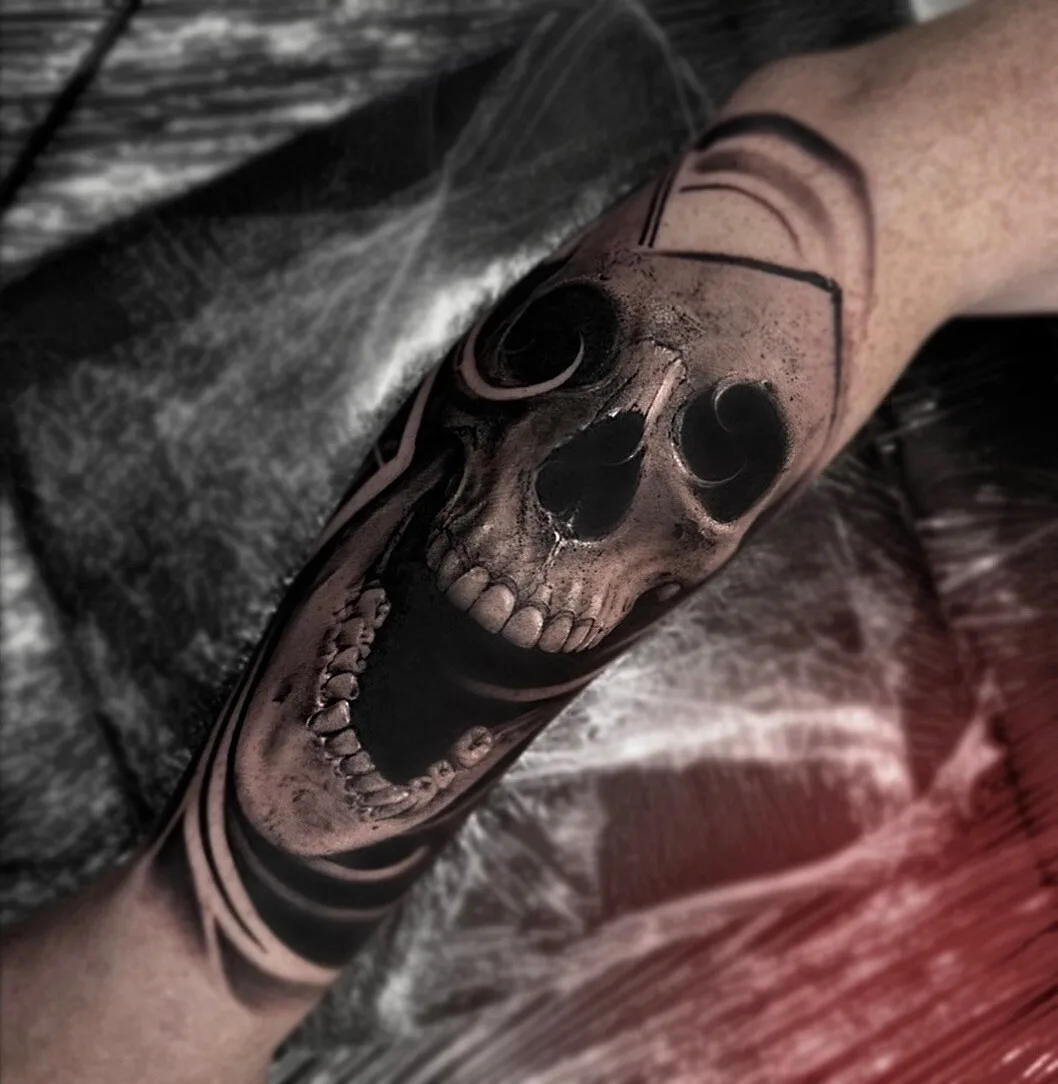 A realistic black and grey skull tattoo placed on the forearm, with intricate details and dark, precise shading.