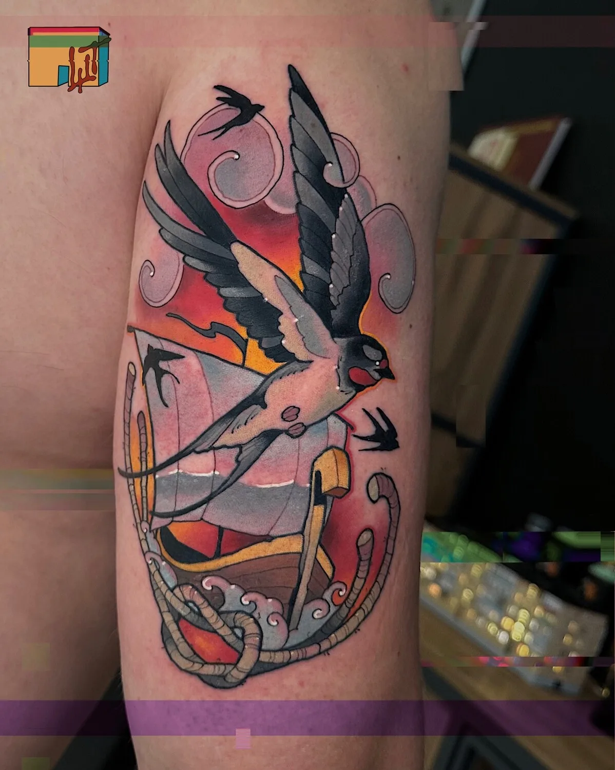 Tatuaggio neo-traditional raffigurante una rondine in volo sopra una barca con sfondo di tramonto colorato.