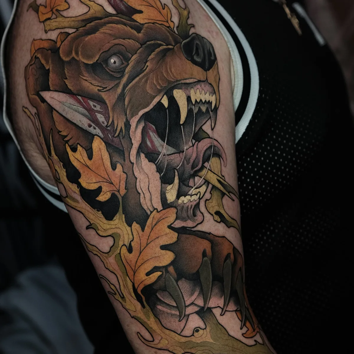 A detailed neo-traditional tattoo featuring a snarling bear with a knife in its mouth surrounded by autumn leaves.