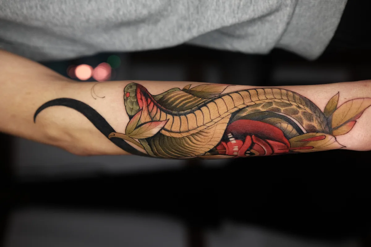 A colourful Japanese-inspired tattoo on the forearm featuring a snake winding through large green leaves, rendered with bold colour and clean outlines.