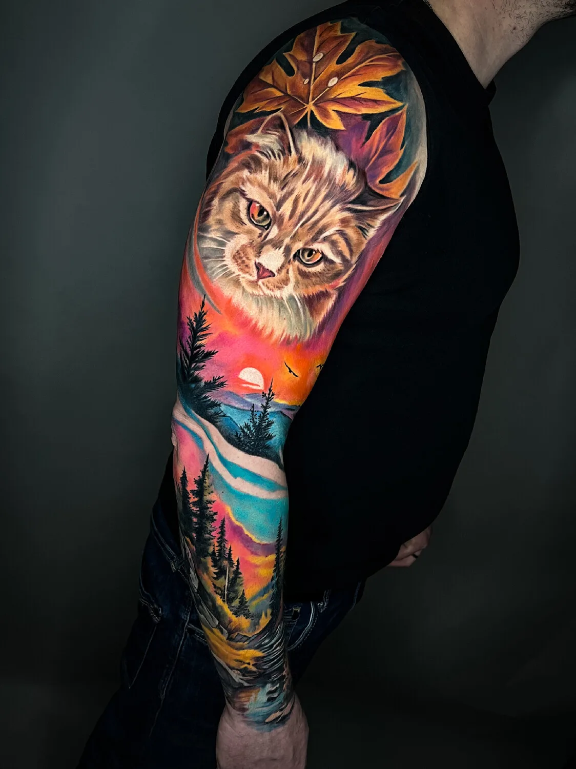 A vibrant full sleeve tattoo combining a realistic cat portrait with a colorful sunset landscape and autumn leaves, covering the entire arm in rich detail.