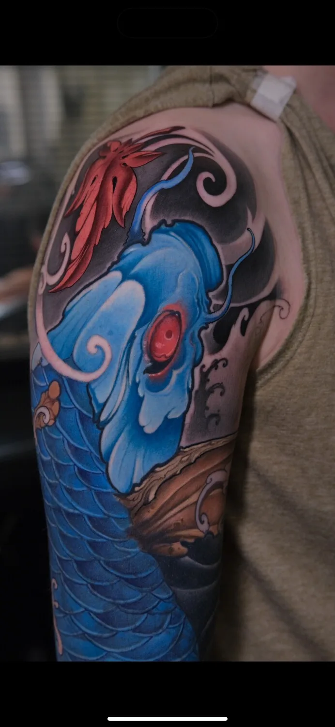 A Japanese-style tattoo on an arm showing a blue koi fish with a red eye alongside a red maple leaf and flowing wave elements.