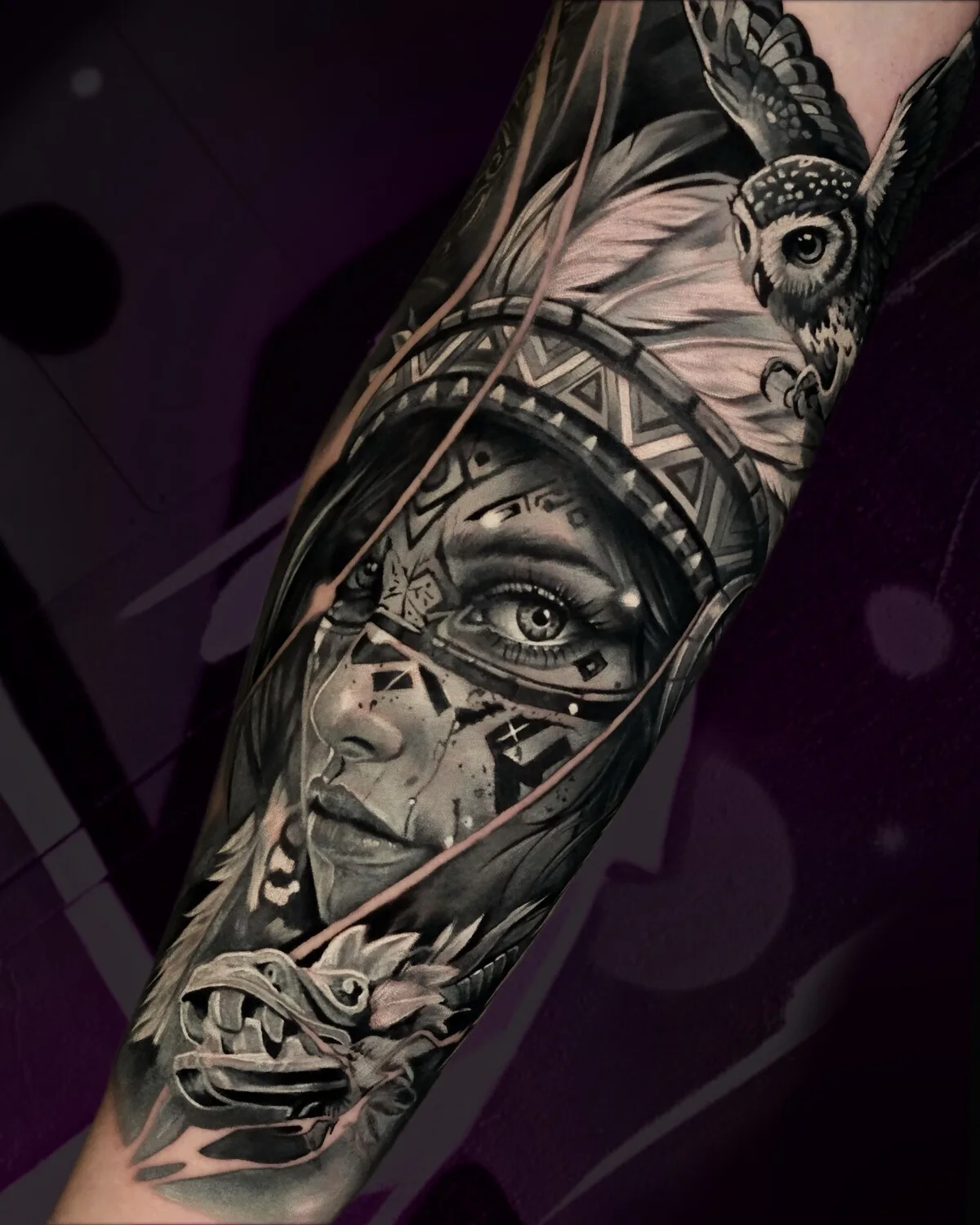 A detailed black and grey tattoo depicting a Native American woman alongside an owl and a dragon-like creature, covering a large area of skin.