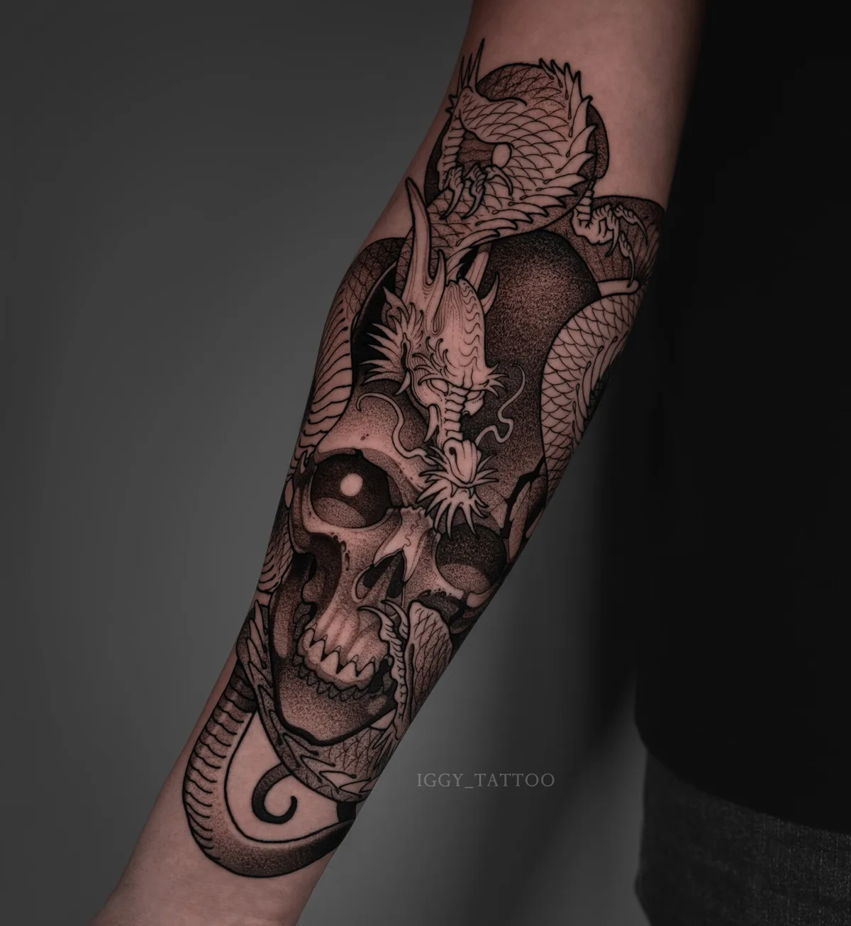 A detailed black and grey tattoo featuring a skull intertwined with a dragon, with intricate linework and shading throughout.