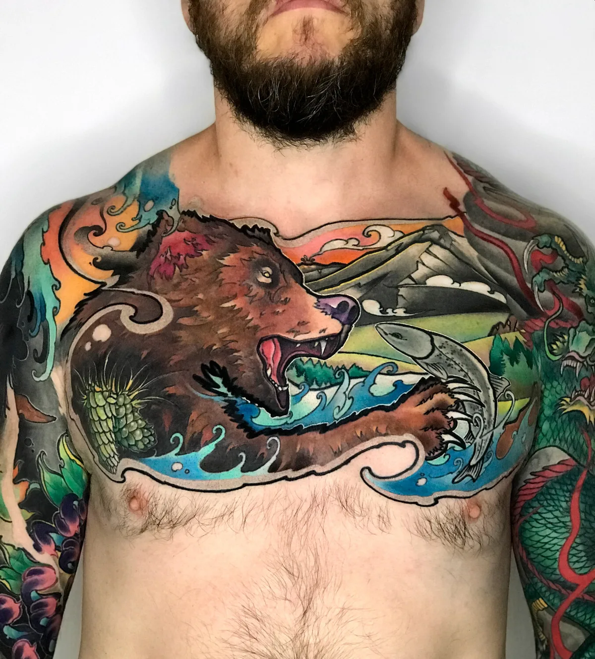 A vibrant Japanese-inspired chest tattoo featuring a bear catching a fish with a landscape background and a dragon on the shoulder.