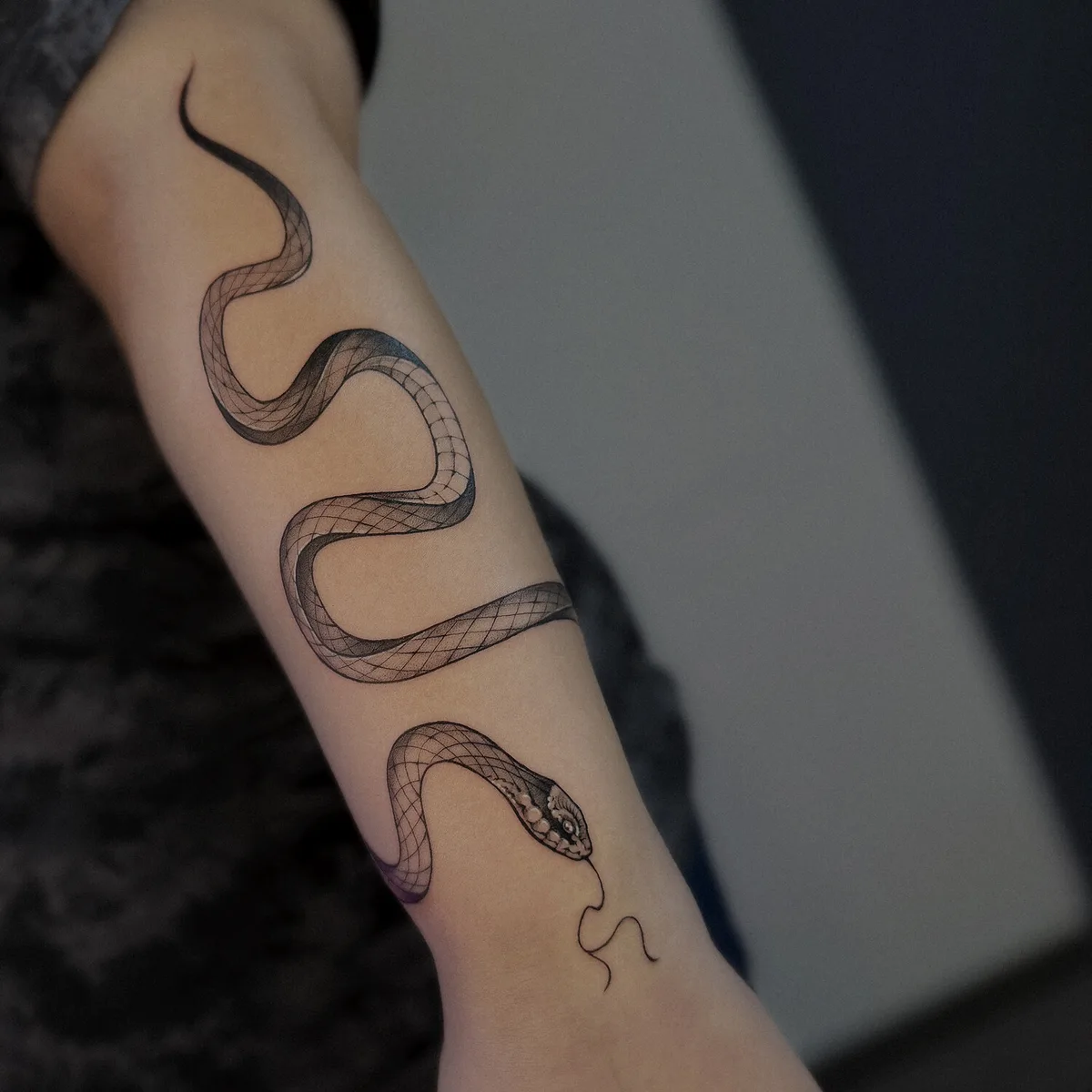 A detailed black and grey snake tattoo wrapping around the forearm, with clean linework and graduated shading showing the coil crossing the joint.