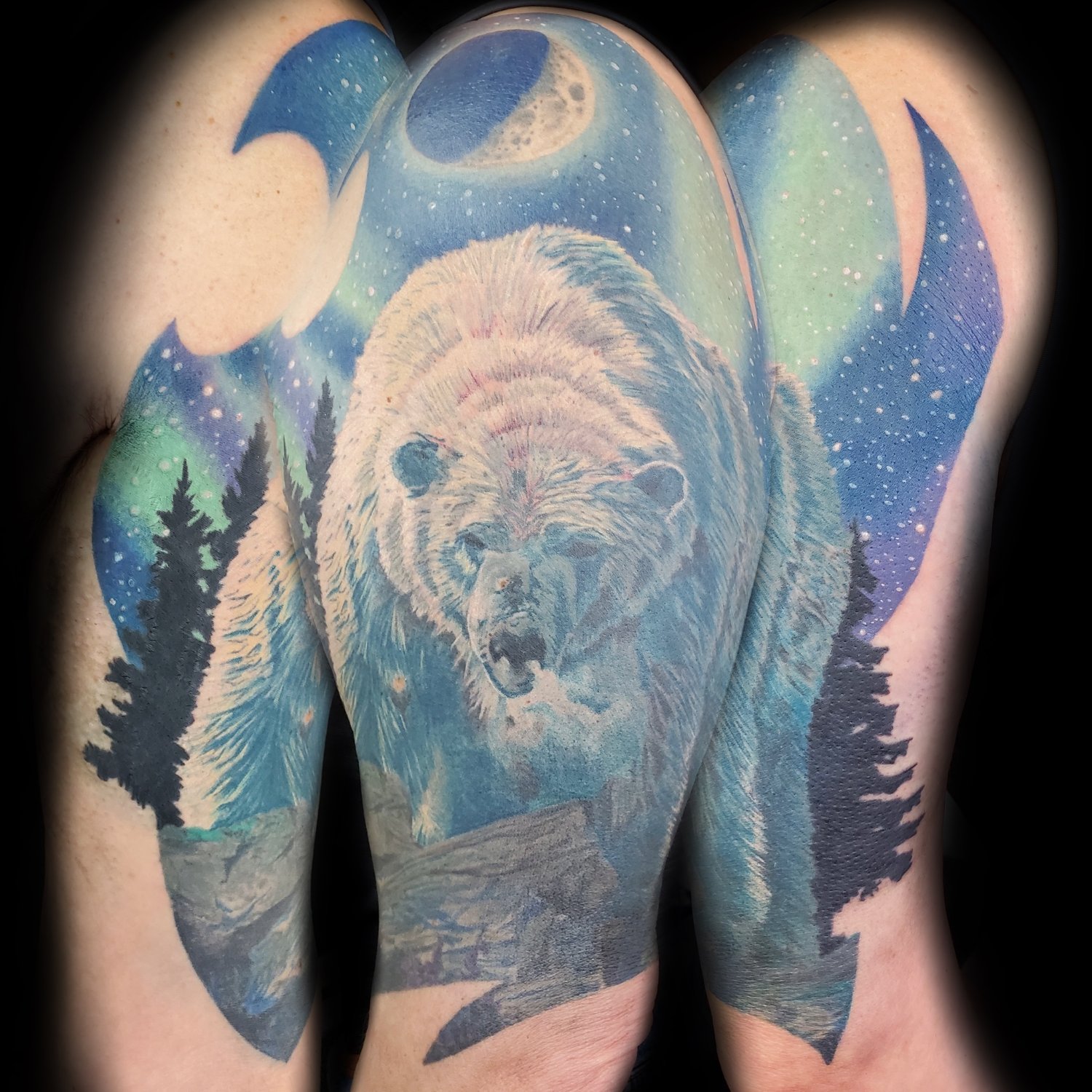 A large illustrative-realism tattoo on an arm and shoulder depicting a polar bear facing forward against a nocturnal Arctic landscape, with a luminous moon, aurora borealis in green and turquoise, and silhouetted pine trees.