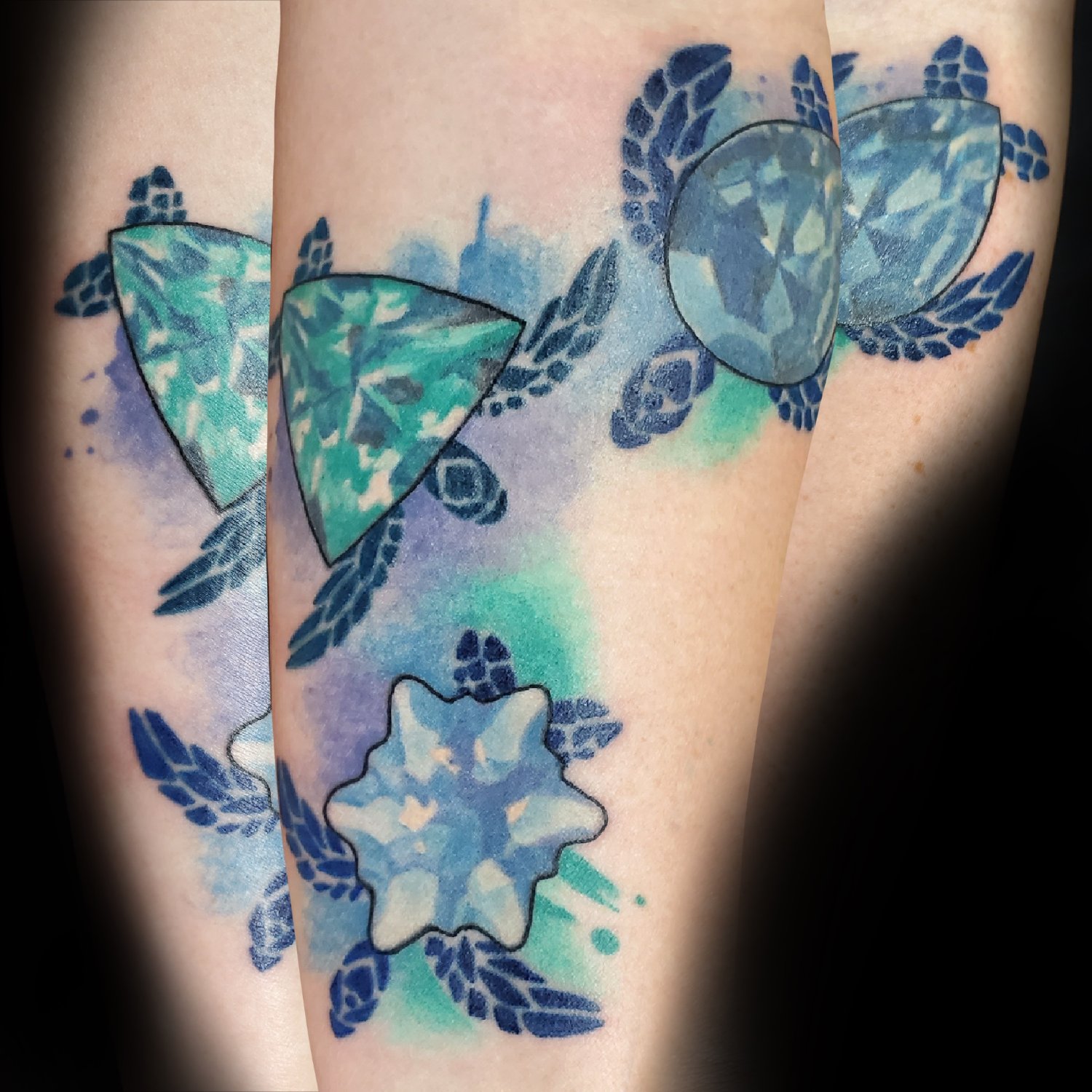 A tattoo on an arm featuring stylised sea turtles with dark blue outlines swimming around faceted gemstones, set against soft watercolour washes of blue, teal, and purple ink.