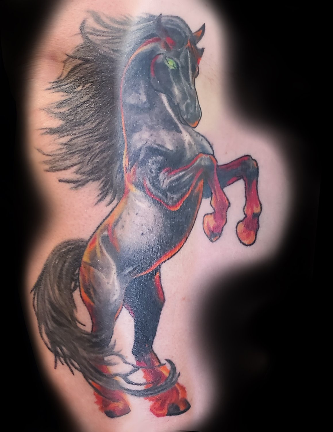A tattoo of a rearing black horse with fiery red and orange accents in its mane and tail and striking green eyes, depicted in a powerful, stylised realistic style with strong shading and bold outlines.