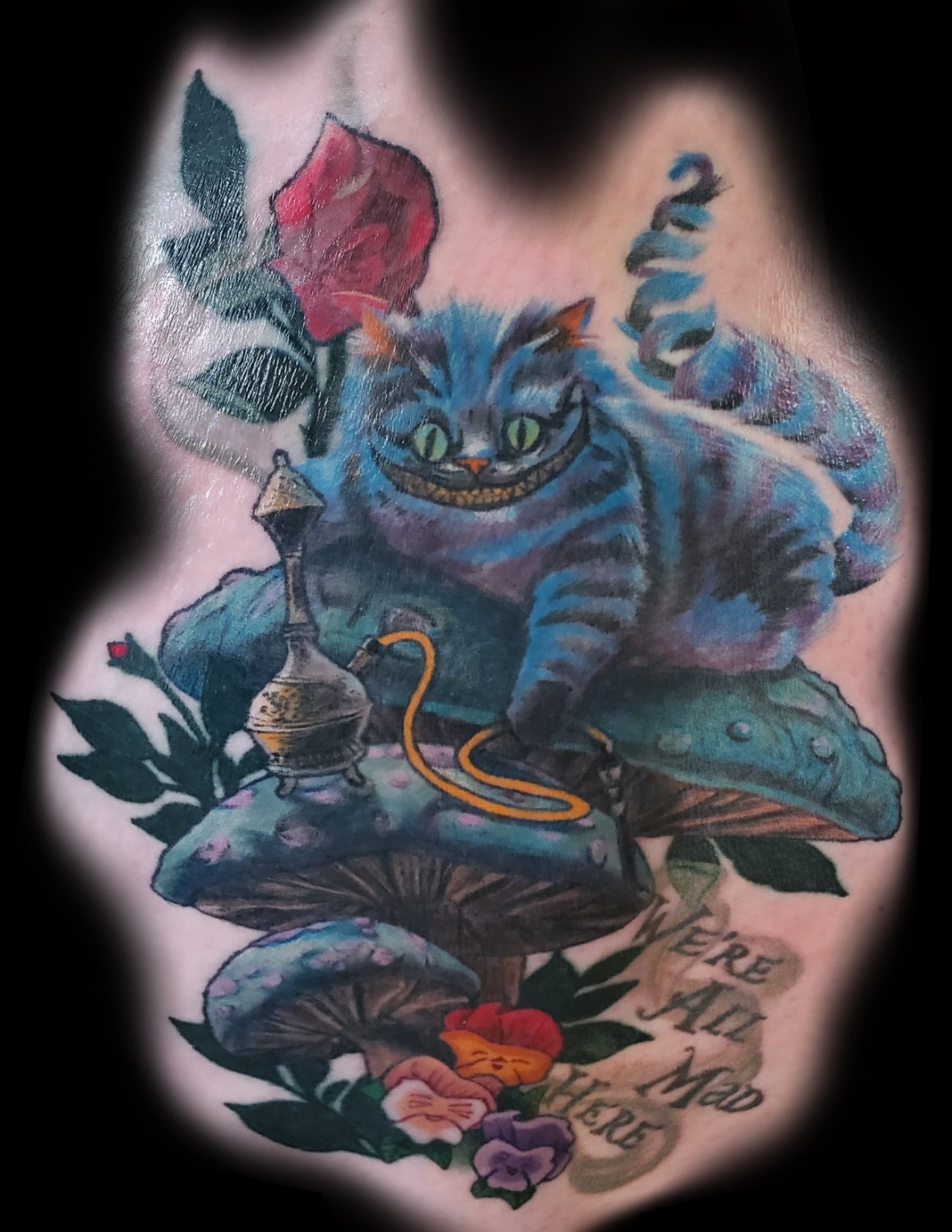 A vibrant illustrative tattoo depicting the Cheshire Cat from Alice in Wonderland, perched on a teal mushroom with a hookah pipe, alongside a rich red rose, smaller mushrooms, colourful pansies, and the text WE'RE ALL MAD HERE in a stylised font.