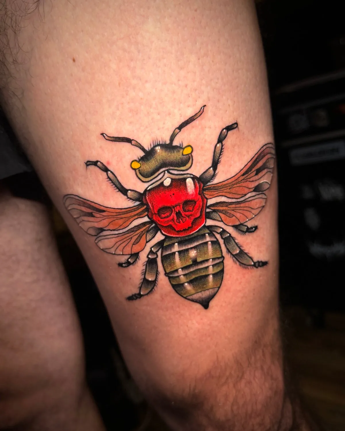 A close-up of a tattoo on pale skin showing a bee viewed from above. Its thorax is a bright red skull with visible eye sockets and teeth. The abdomen is olive green with dark stripes, the wings are translucent amber-brown, and thick black outlines define every element throughout.