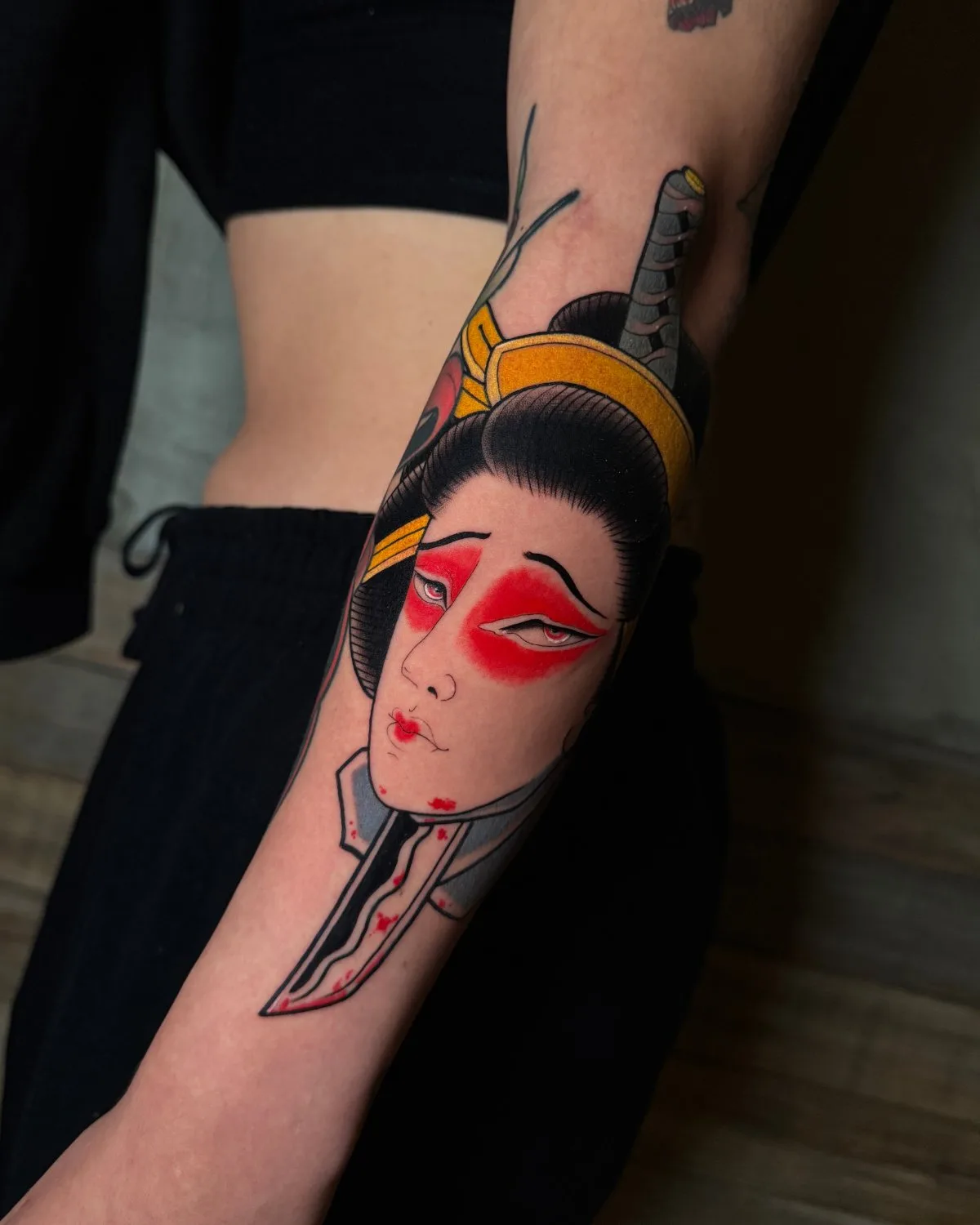A forearm tattoo in neo-traditional style showing a geisha's face and shoulders. She has bold red eyeshadow, deep red lips, and dark hair in a bun with a gold headband. A katana runs vertically alongside her face, its blade marked with blood. Bold black outlines and saturated flat color define the p