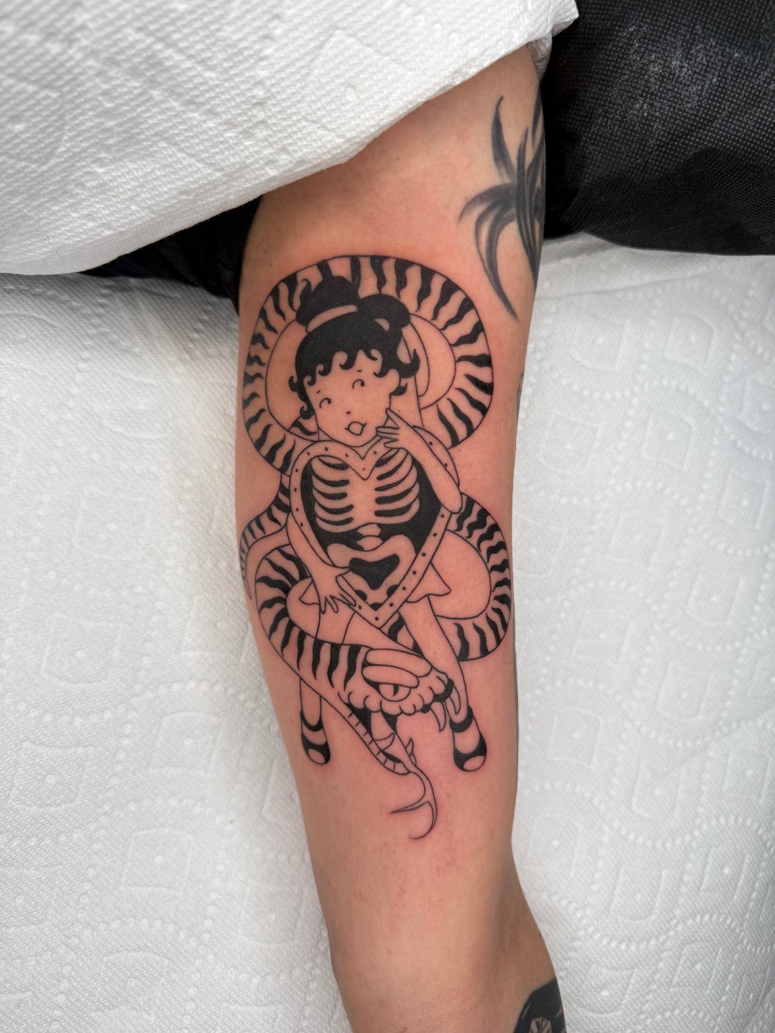 A black ink tattoo on an arm showing a round-faced child figure with curly hair whose torso is rendered as a visible ribcage. A bold black-and-white striped snake coils around the child and the heart-shaped ribcage, its open mouth and forked tongue at the bottom of the design.