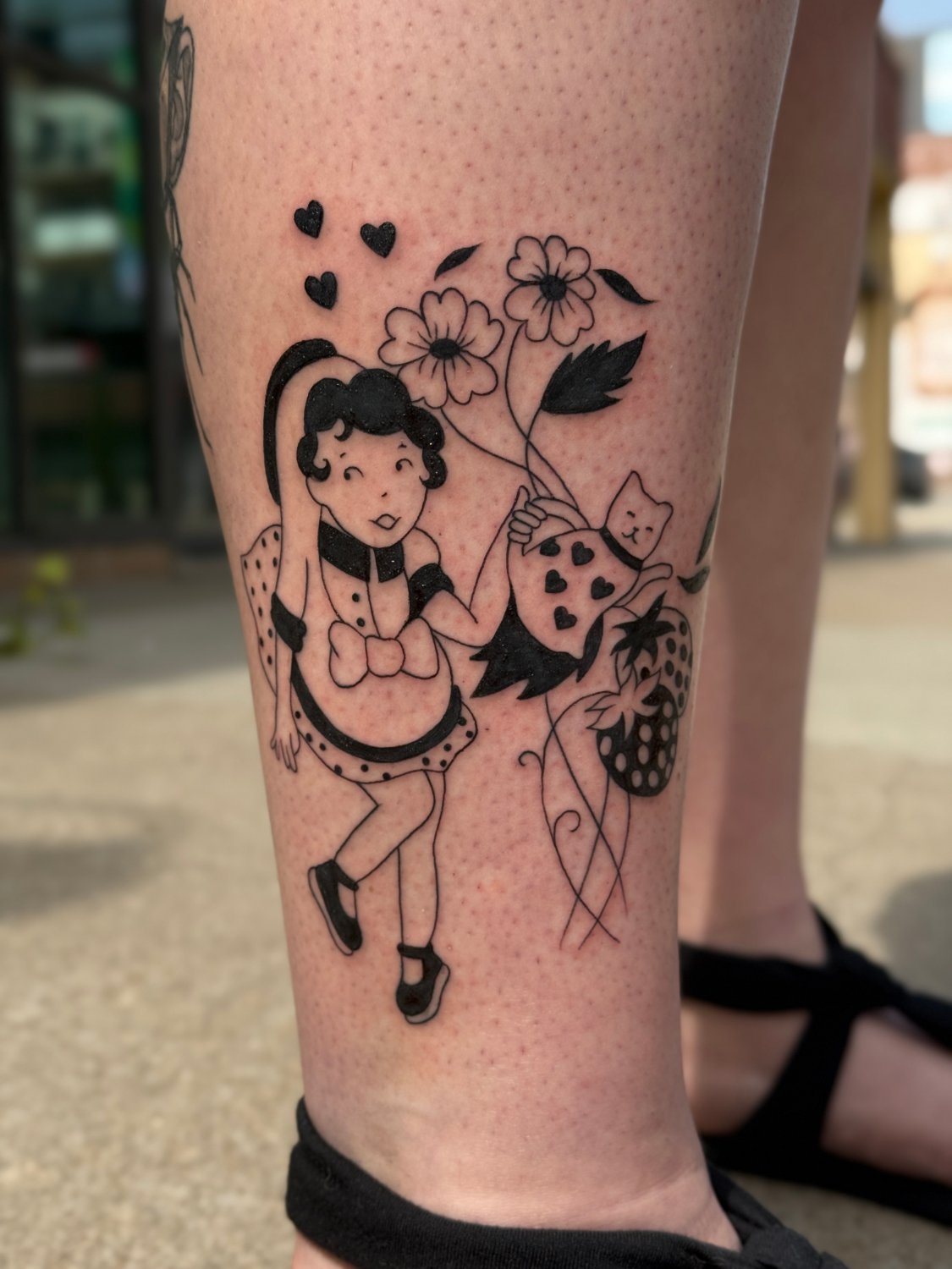 A black ink tattoo on a leg depicting a cheerful girl in a polka-dot dress with a bow at the neck, holding a bouquet of stylised flowers and a heart-decorated bag. A small cat peeks from behind the bouquet. Three solid black hearts float above her head. A large strawberry with curling vines sits to 