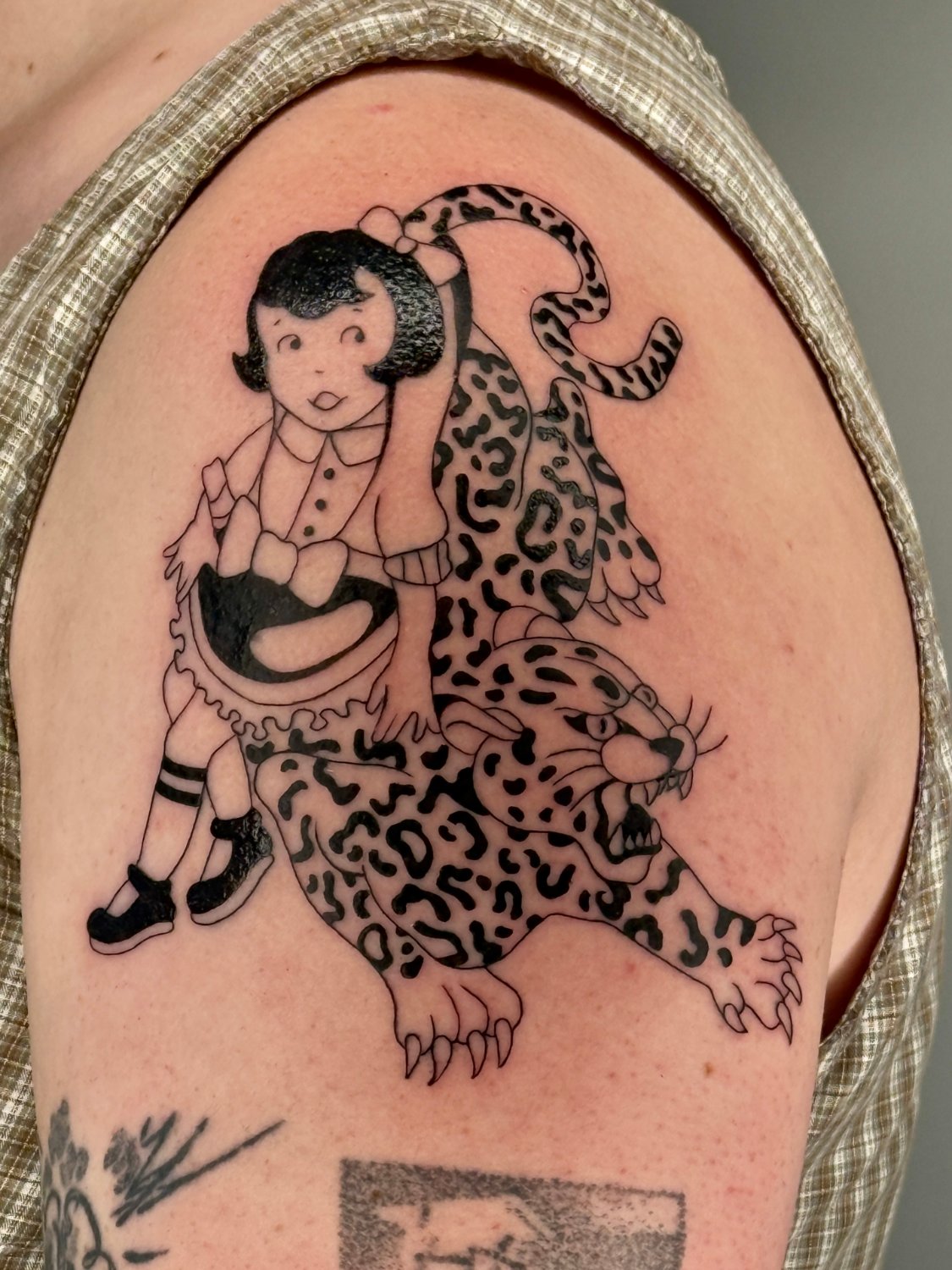 A black ink tattoo on the upper arm of a girl in a ruffled-collar apron dress with a bow in her hair, seated upright on the back of a snarling leopard with open mouth, extended claws, and a long spotted tail curling upward behind her.