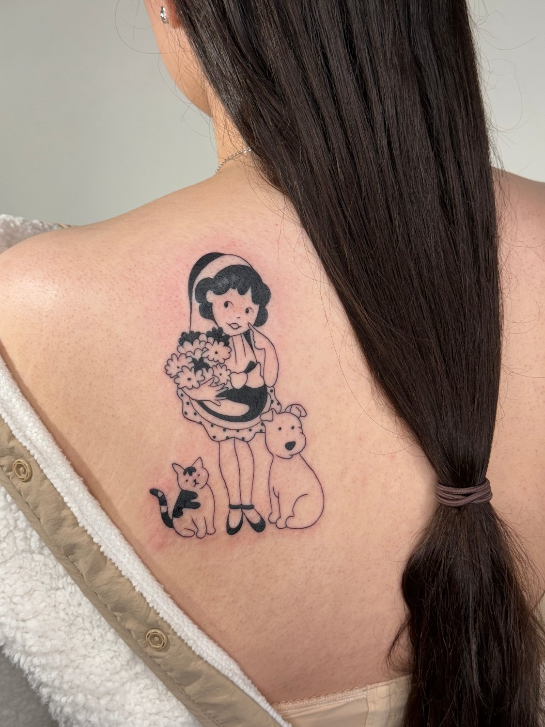 A black ink tattoo on the upper back showing a curly-haired girl with a headband and finger near her mouth, holding a bouquet of round flowers. A black-and-white cat sits to her left and a light-coloured dog sits to her right. The design is in clean illustrative linework with minimal shading.