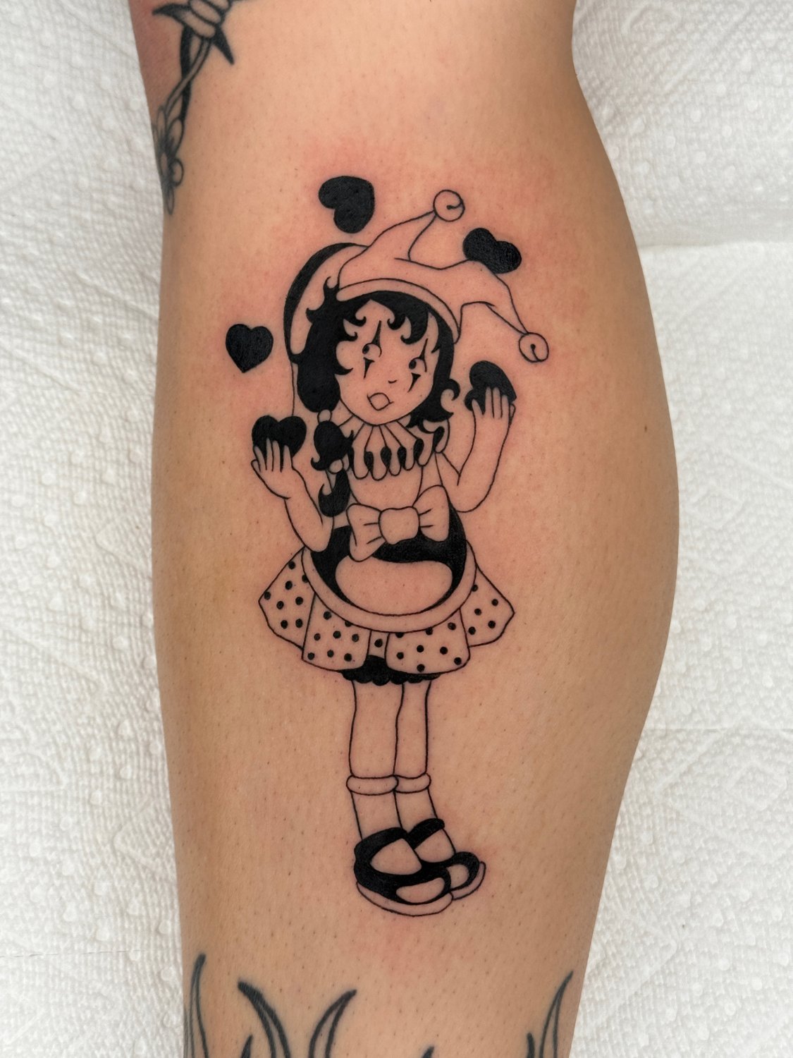 A black ink tattoo on a leg showing a jester-like clown girl in a floppy hat with heart-tipped points, a ruffled collar, polka-dot skirt, and sandals. She juggles three solid black hearts, with two more floating near her head. Another tattoo of a dagger is partially visible above.