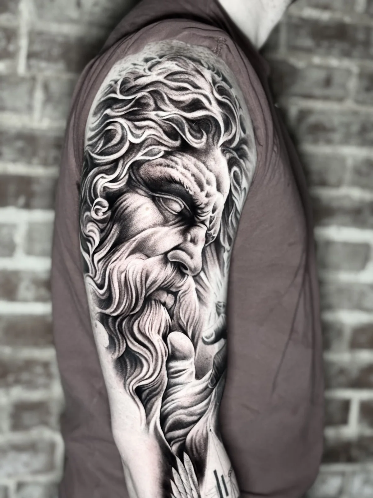 Black and grey realism tattoo of a bearded Zeus-like god on the upper arm