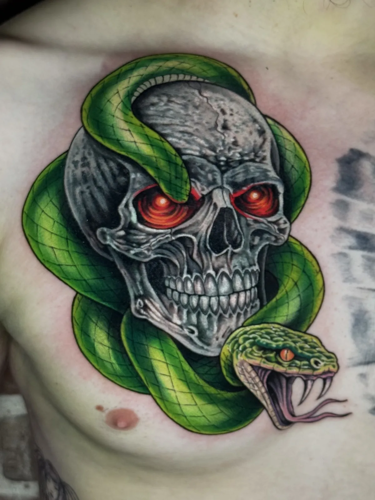 Color realism chest tattoo of a chrome skull with glowing red eyes and a green snake coiled around it