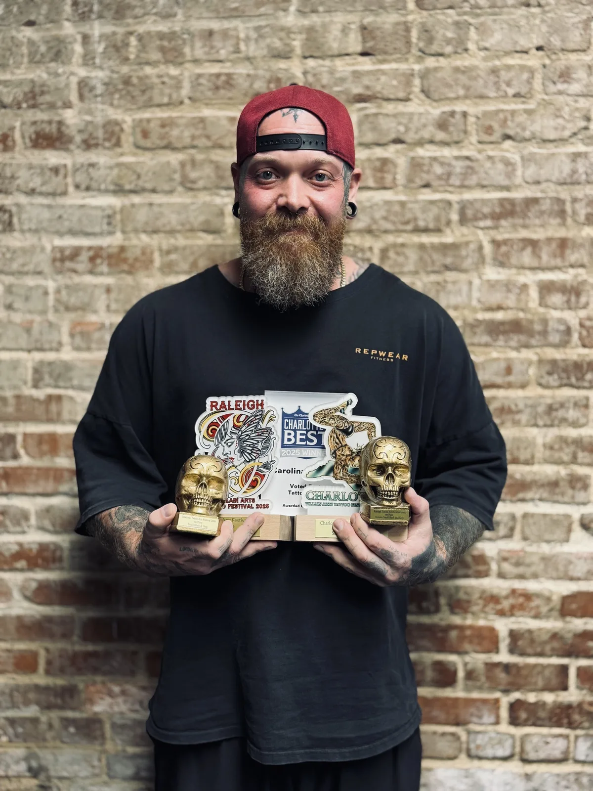 Eike Schumacher holding convention trophies and the Charlotte's Best 2025 second-place award for Best Tattoo Shop