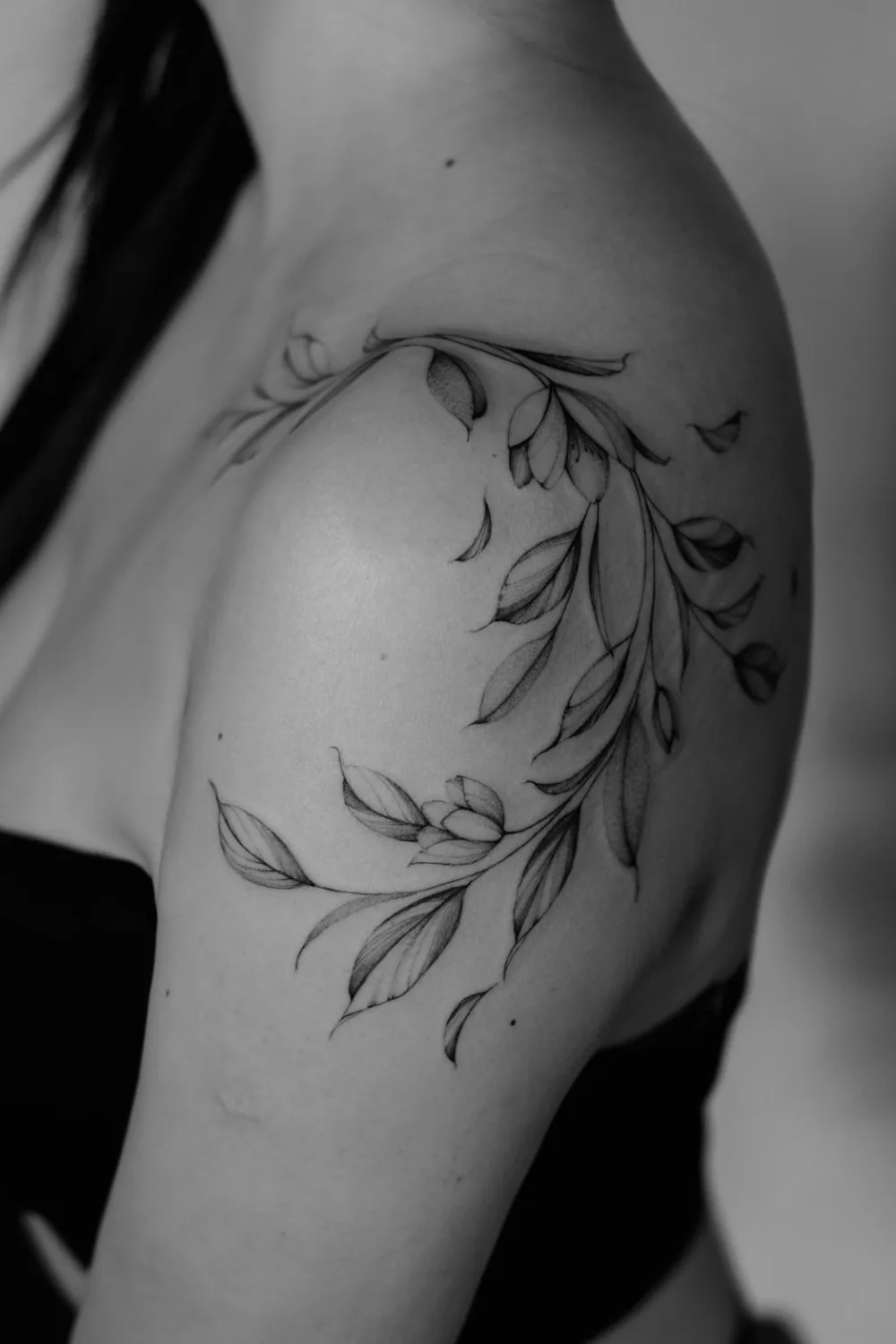 Black and white photograph of a shoulder and upper arm bearing a fine-line tattoo of a curving vine with small leaves and buds, some leaves appearing to float free of the main stem.
