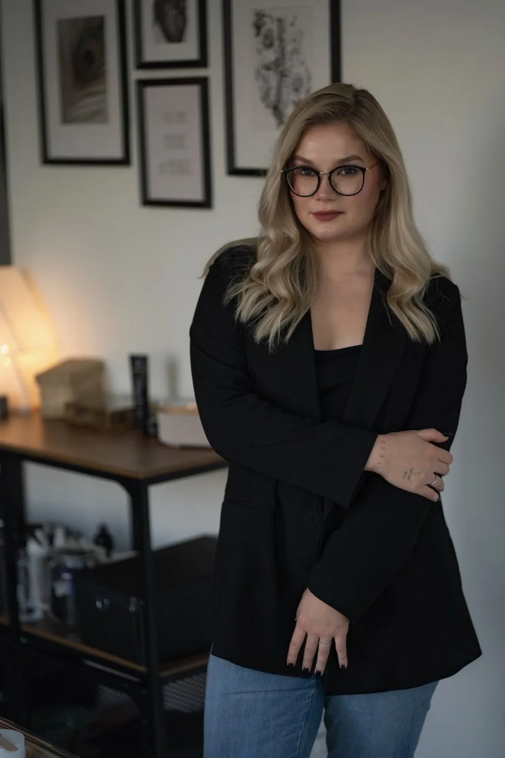 A blonde woman with glasses in a black blazer stands with arms crossed in front of a white wall hung with three framed prints including a peacock feather, a quote, and an anatomical drawing.