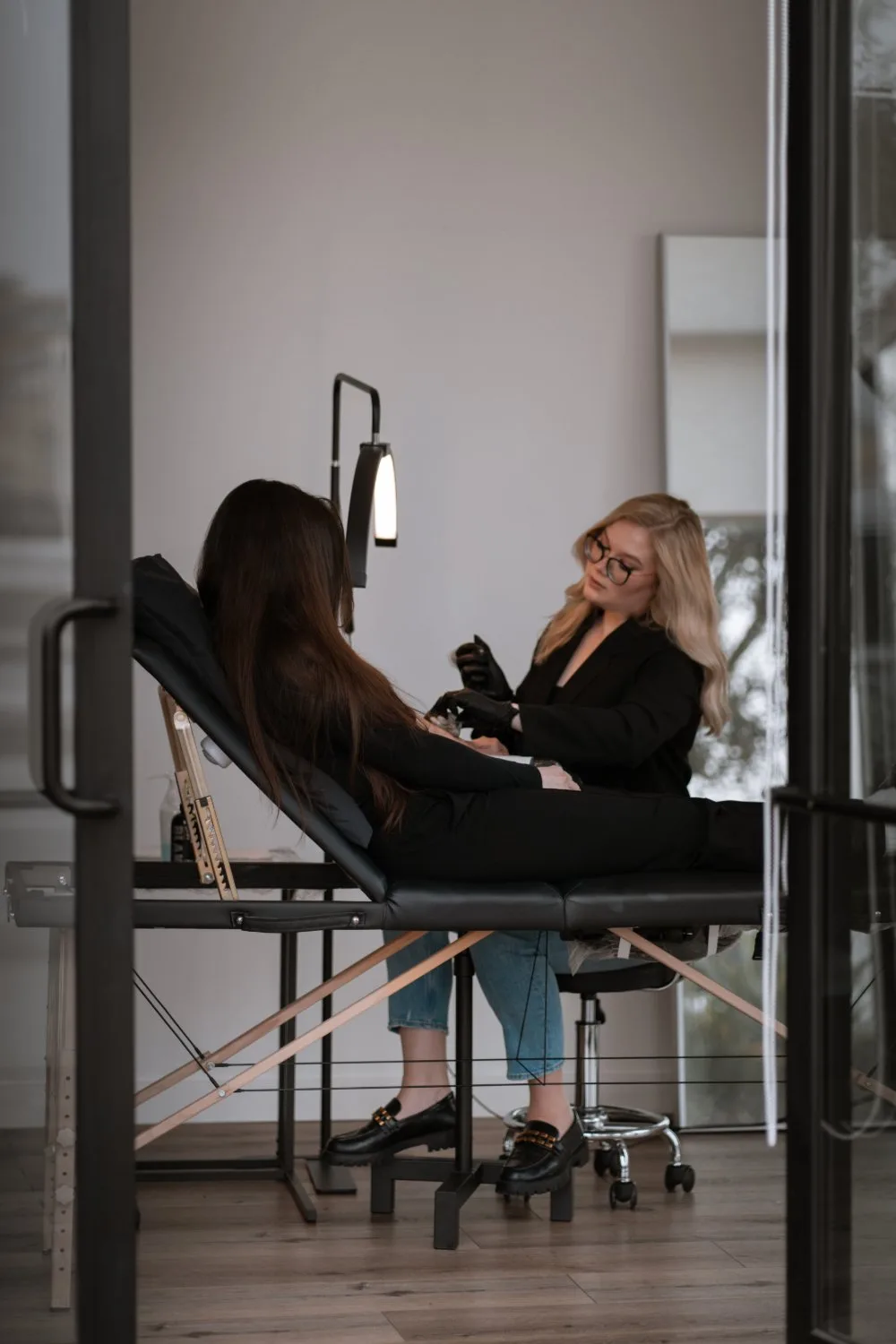A blonde woman with glasses leans over a client reclined on a black treatment bed in a modern studio, a bright adjustable lamp overhead, the scene viewed through a doorway.