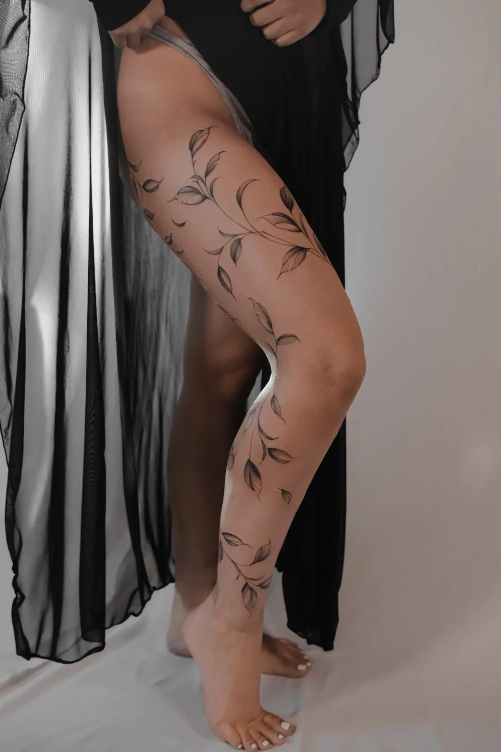 A woman's legs showing a black fine-line tattoo of a vine with numerous leaves curling from calf to upper thigh, the design leaving generous negative space against pale skin.