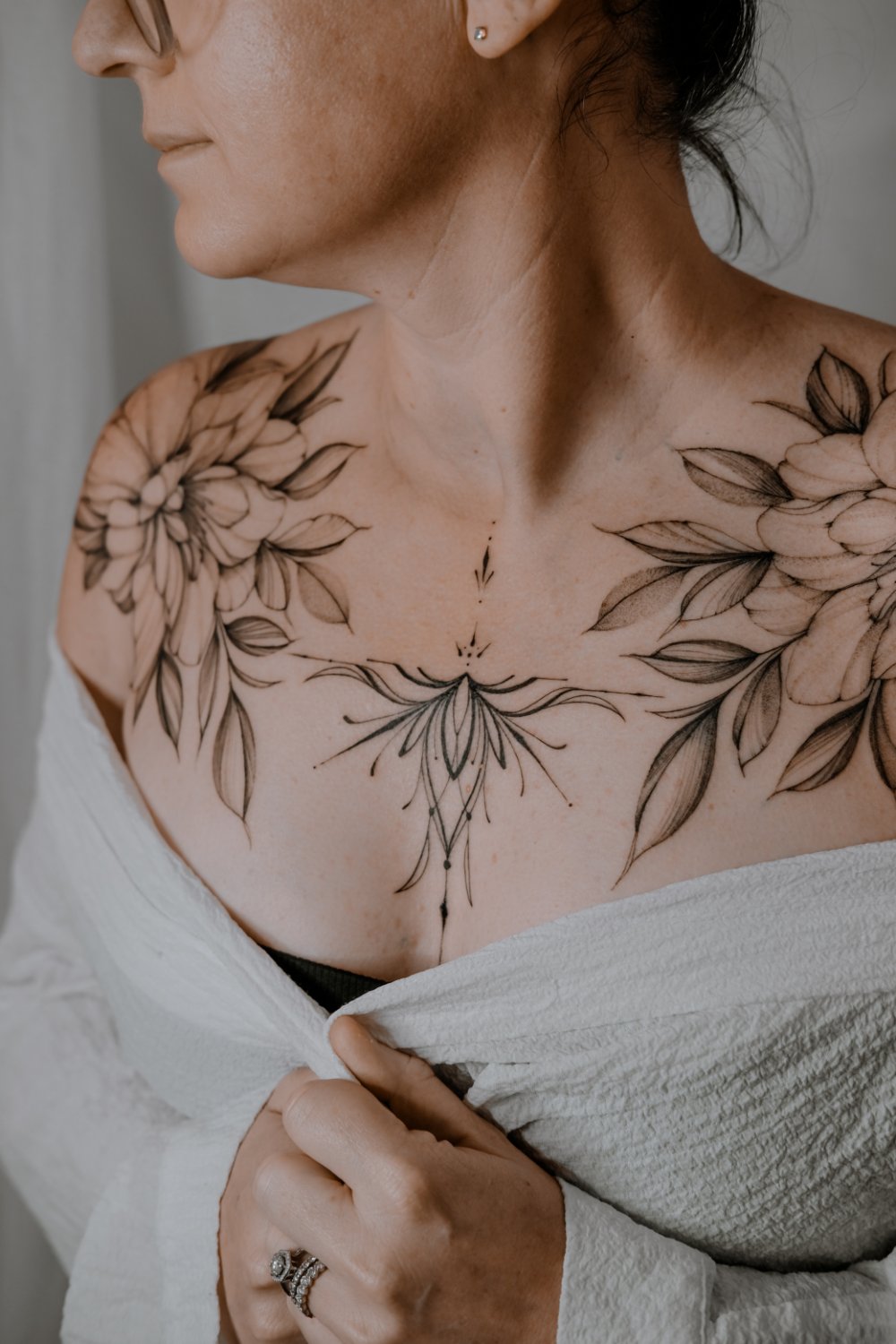 A woman's chest and shoulders showing a large fine-line tattoo of floral motifs on each shoulder connected by a central lotus-like design with radiating lines and dot details at the sternum.