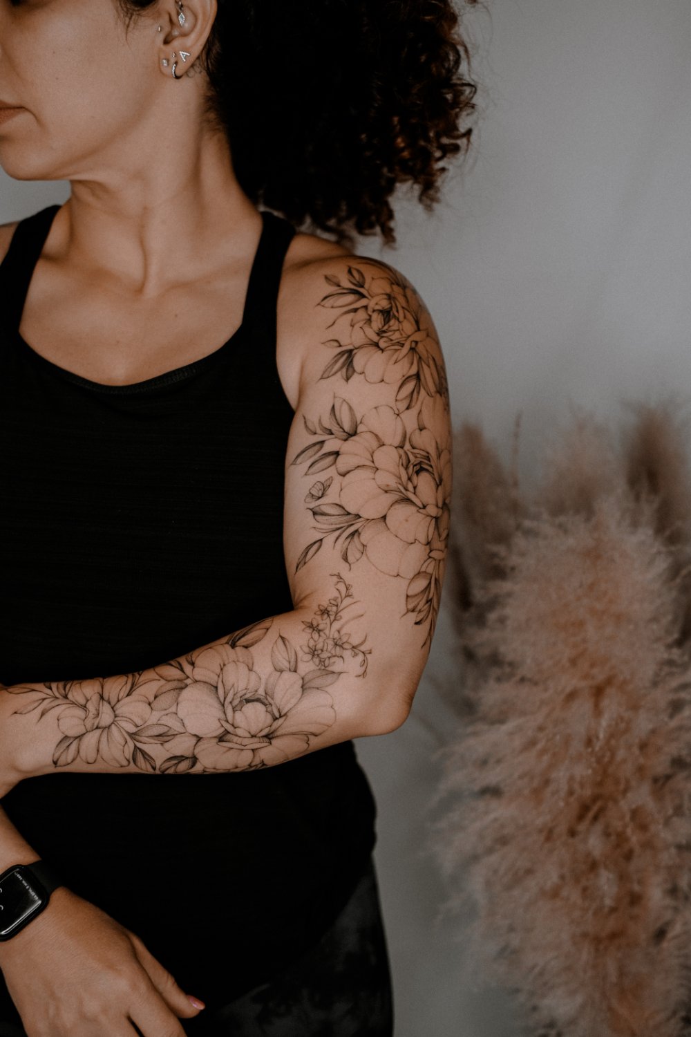 A woman in profile showing a full-sleeve black fine-line tattoo of large open peonies, smaller blossom clusters, leaves, and a small butterfly covering the arm from shoulder to forearm.