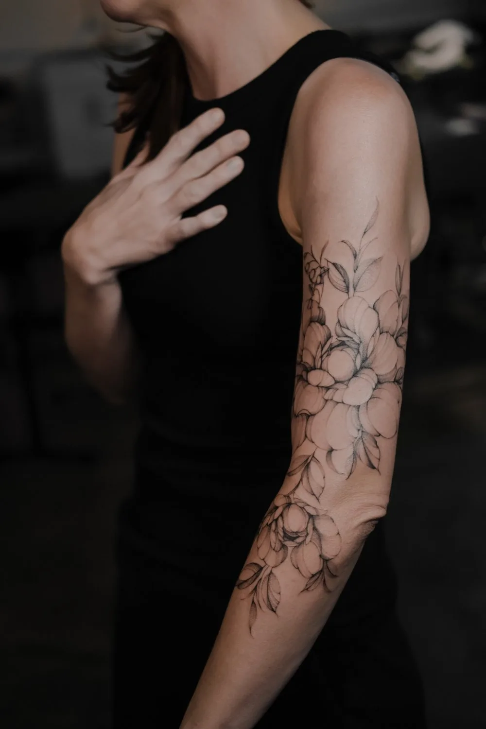 A woman's upper arm and shoulder showing a black and grey fine-line tattoo of large peony blooms, buds, and elongated leaves arranged in a cascading composition that follows the curve of the arm.