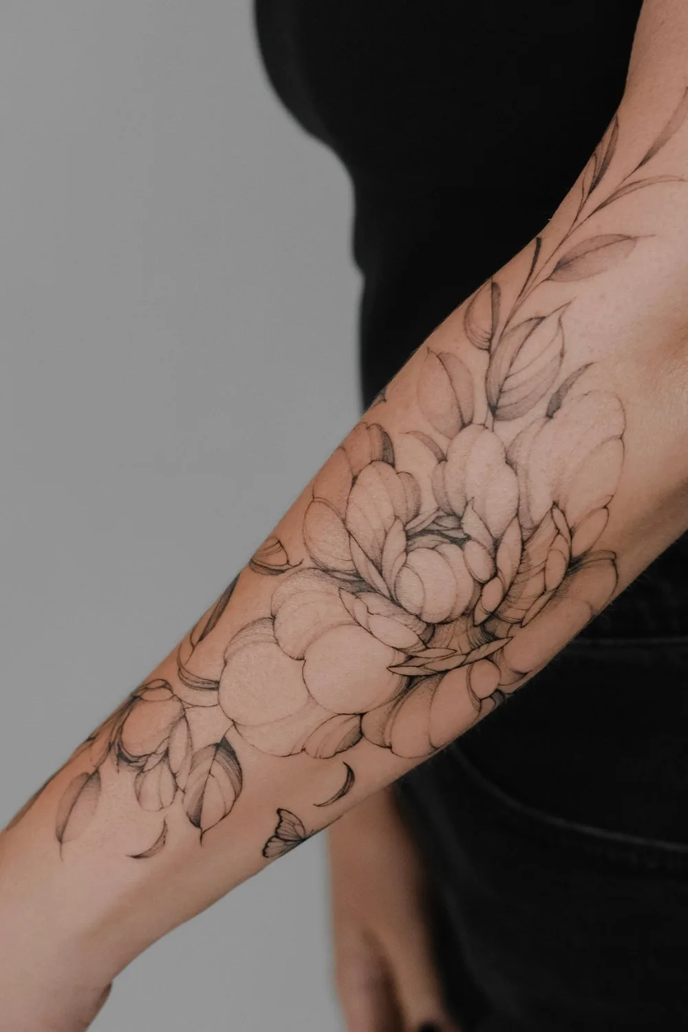A person's forearm showing a black fine-line tattoo of a large multi-petalled flower, a branch with leaves extending toward the elbow, and a small stylised butterfly near the lower forearm.