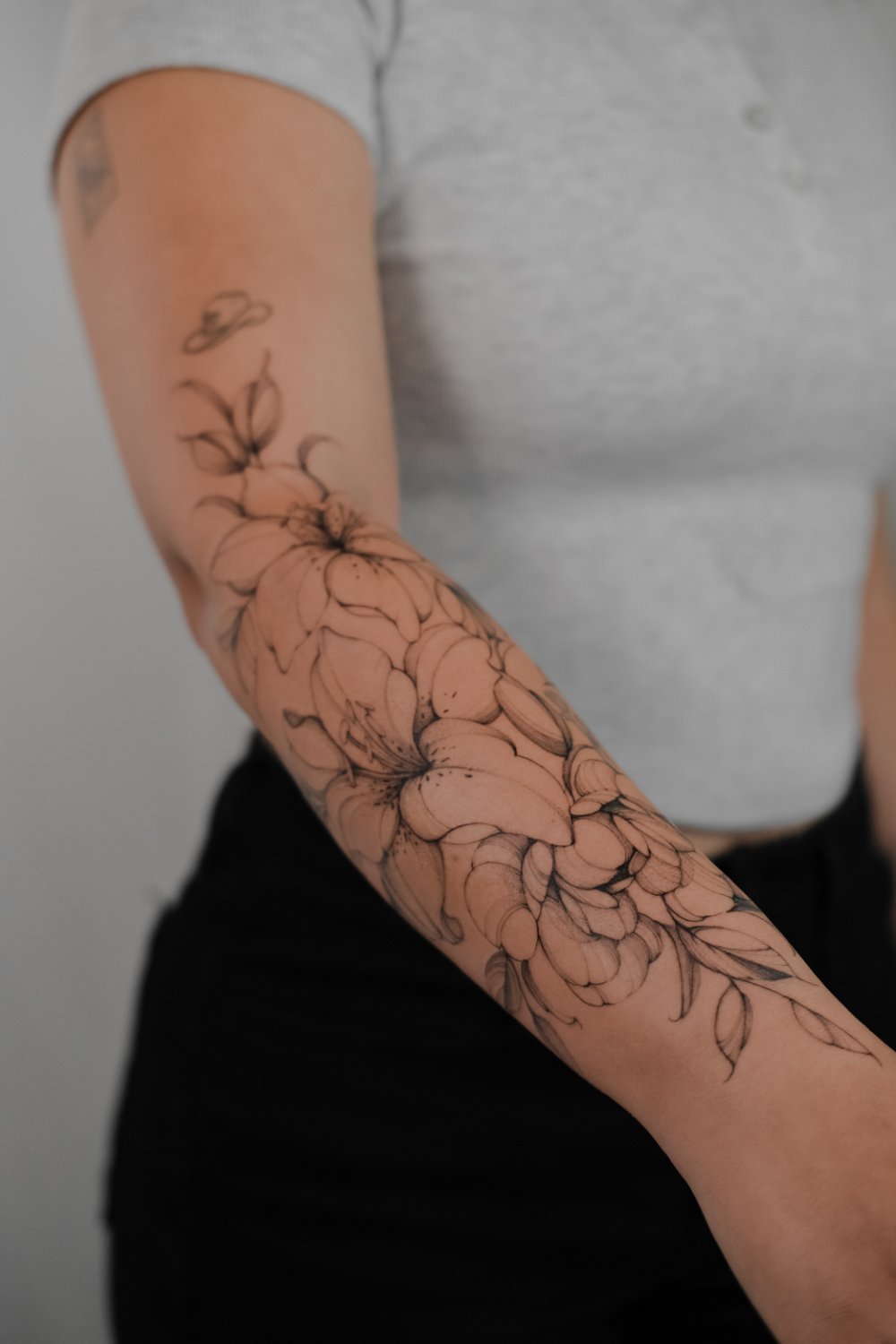 A person's forearm and upper arm bearing a fine-line botanical tattoo of lily-like flowers with prominent stamens, layered peony-style blooms, elongated leaves, and smaller abstract elements near the shoulder.