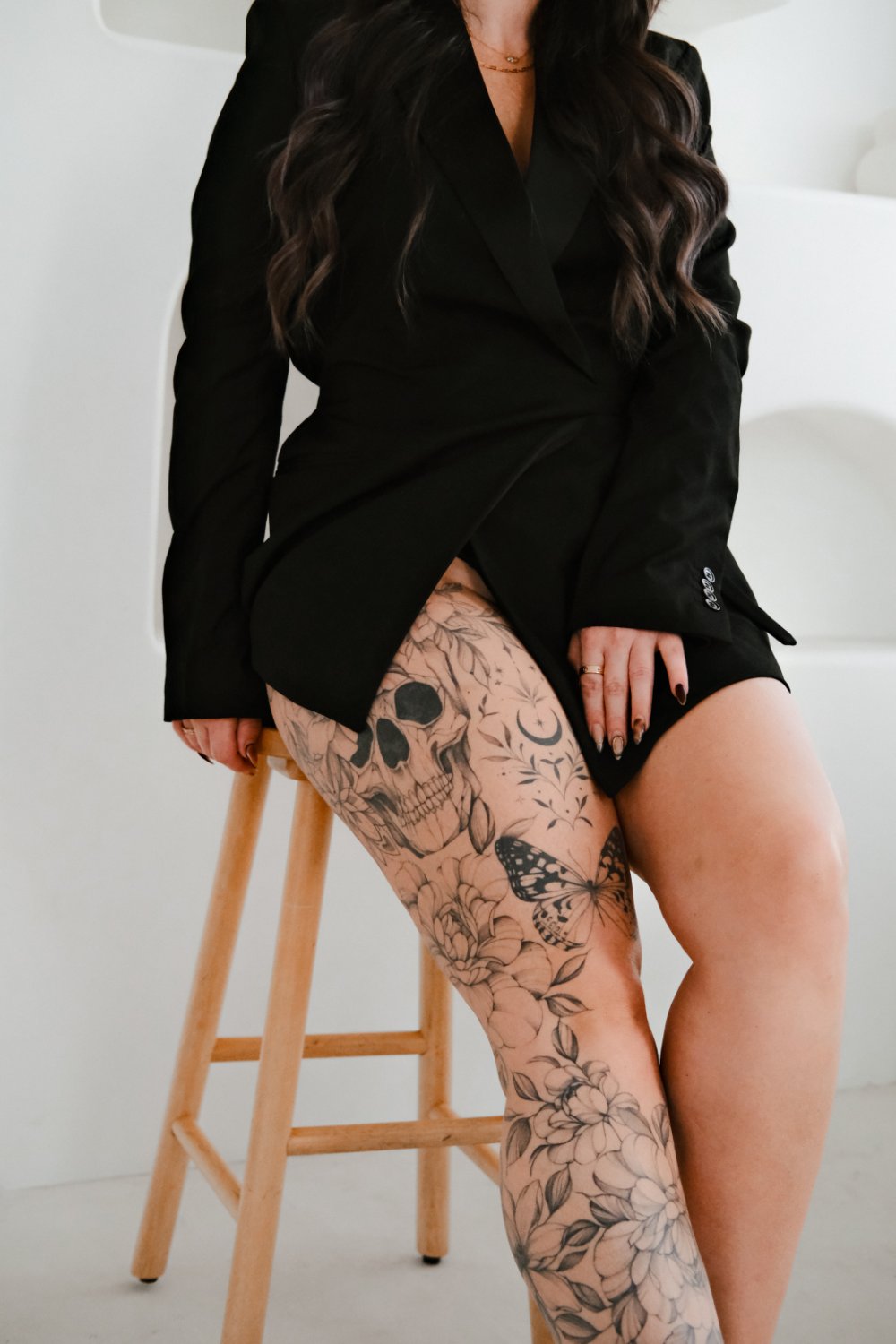 A woman seated on a wooden stool with both legs visible, showing detailed black and grey tattoos including a realistic skull, fine-line floral patterns, a crescent moon, and a butterfly spread across the thighs and lower legs.