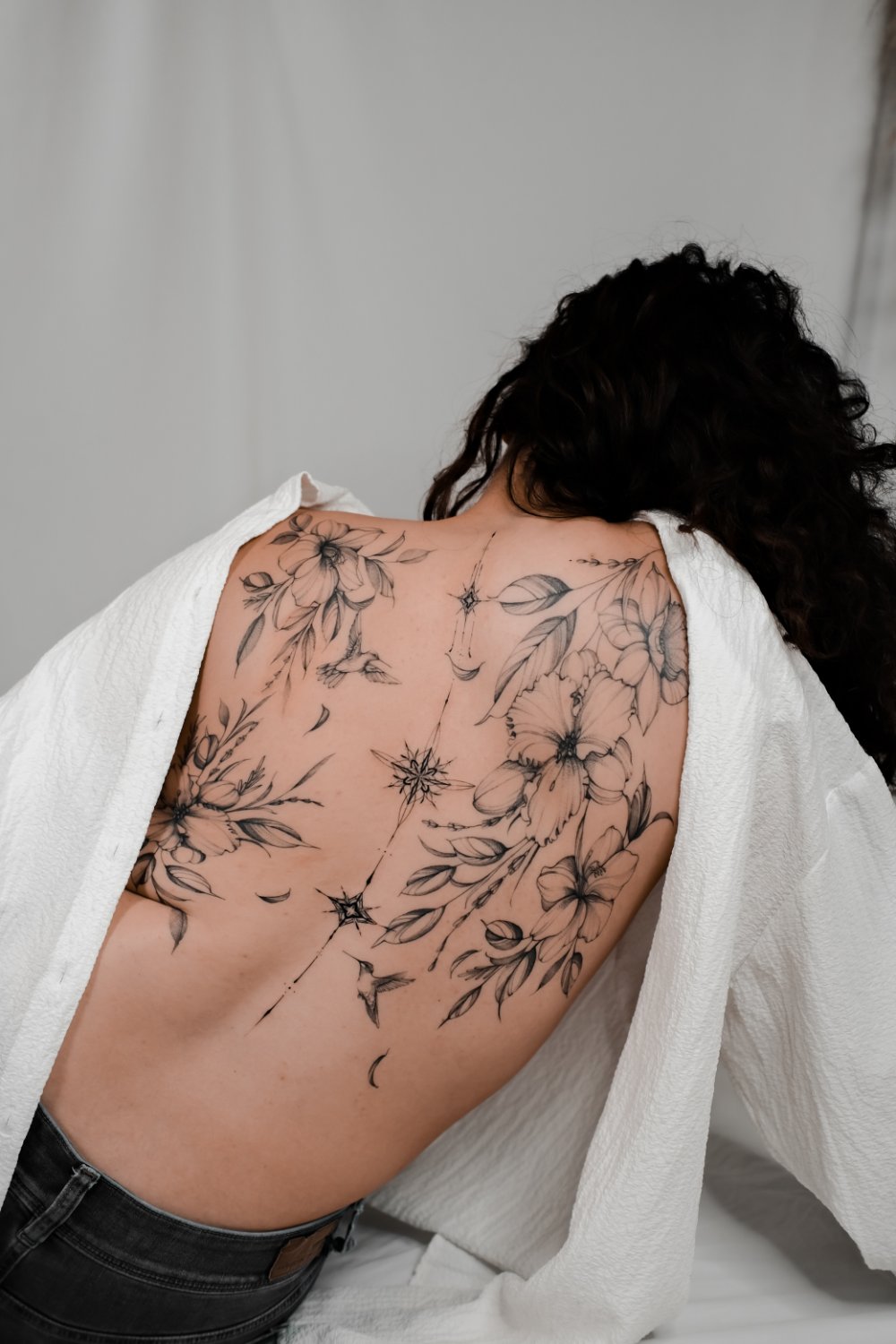 A person's full upper back covered by a fine-line black and grey tattoo of open lily and hibiscus flowers, broad and slender leaves, small hummingbirds in flight, and celestial compass-rose and crescent-moon symbols connected by thin lines along the spine.
