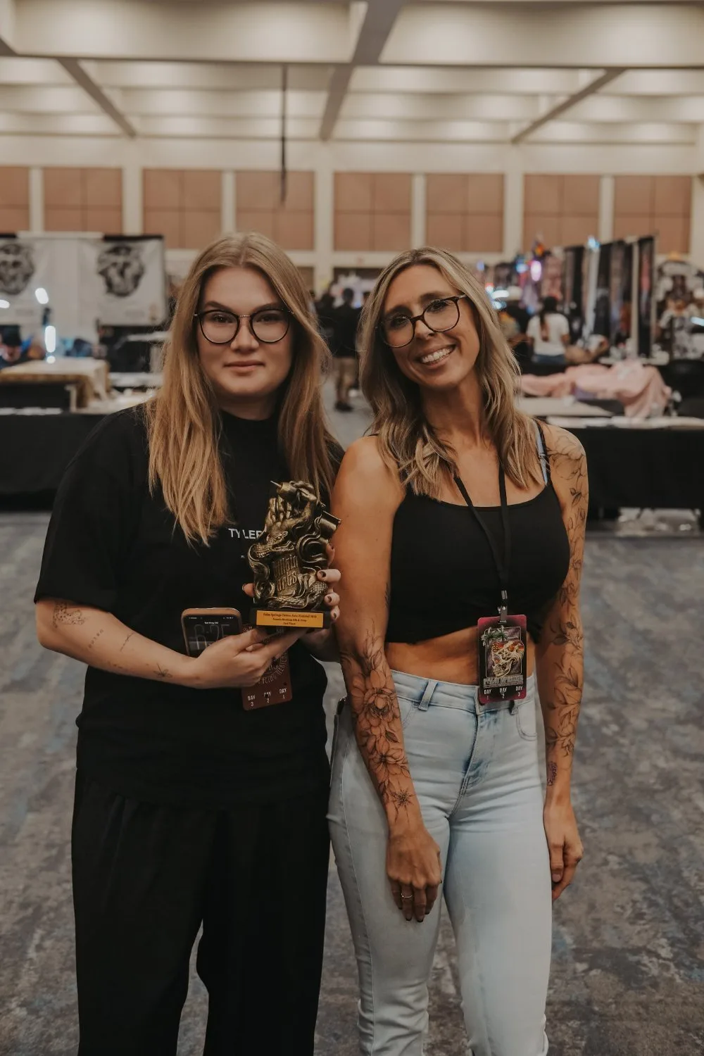 Two blonde women with glasses stand side by side indoors at a convention; the woman on the left holds a bronze sculptural trophy while the woman on the right smiles, her arms showing floral tattoos.