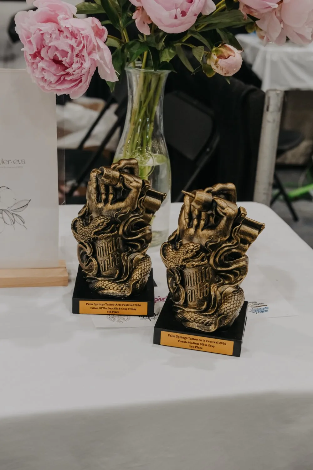 Two bronze hand-shaped trophies on black bases, their plaques inscribed with Palm Springs Tattoo Arts Festival 2026 placing results, set beside a vase of pink peonies on a white tablecloth.