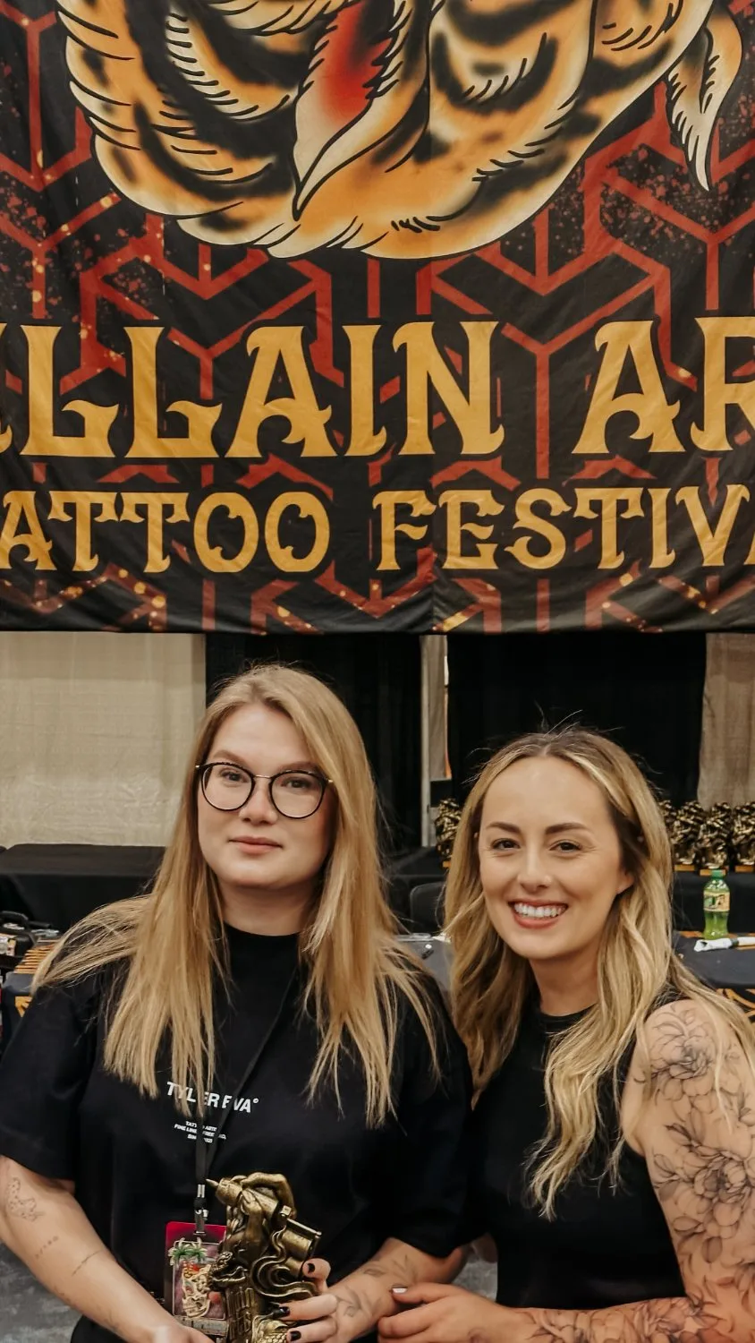 Two blonde women stand before a Villain Arts Tattoo Festival banner; the woman on the left holds a gold trophy and wears a Tyler Eva branded t-shirt, the woman on the right displays detailed floral arm tattoos.