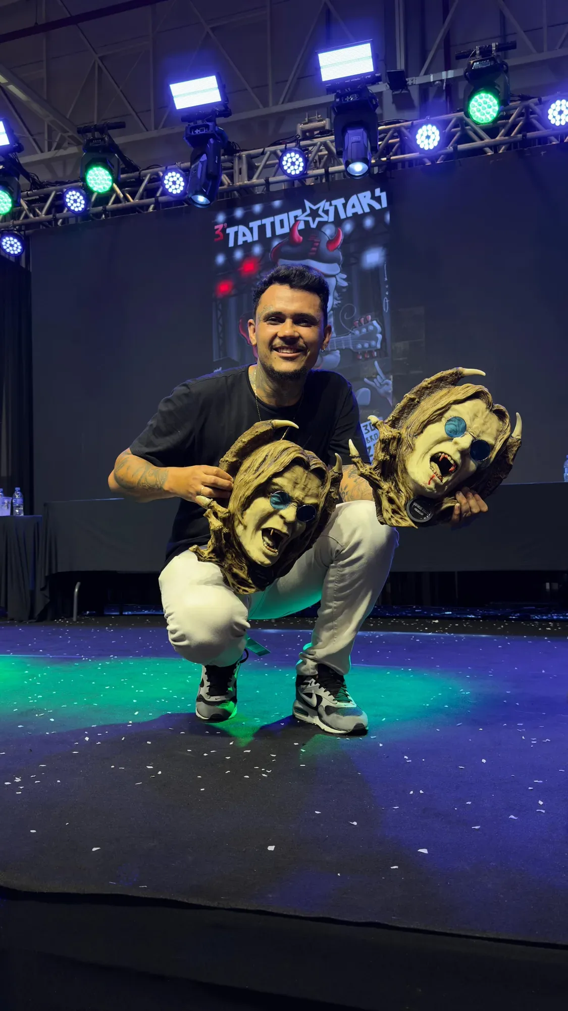 A man with dark hair and a beard crouches on a lit stage holding two pale grotesque mask-shaped trophies, one in each hand; green and blue stage lights illuminate the floor and a large screen behind him reads '3 TATTOOSTAR'.