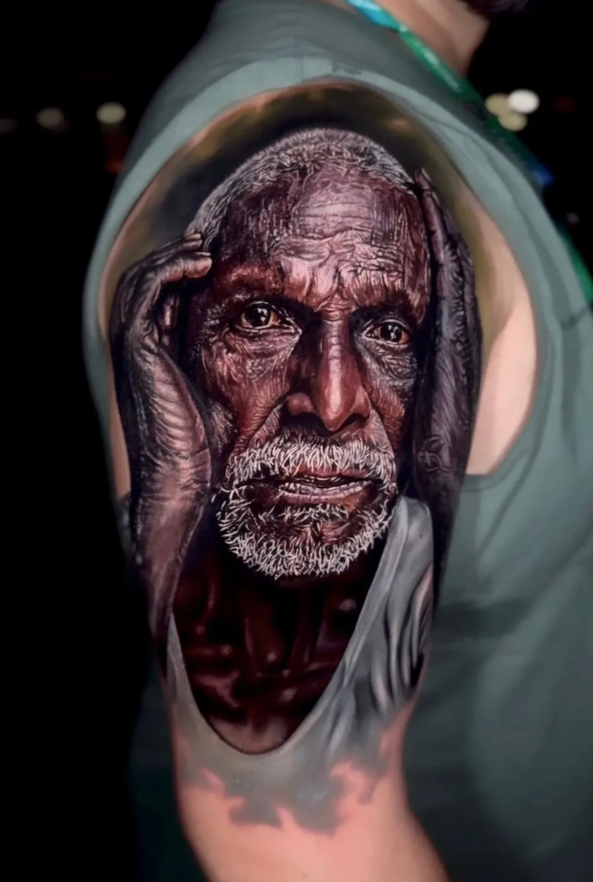 A hyperrealistic tattoo of an aged man's face with a white beard on the upper arm and shoulder; the man's hands frame his face and the lower edge of the tattoo creates the illusion of skin being peeled back to reveal the portrait beneath.