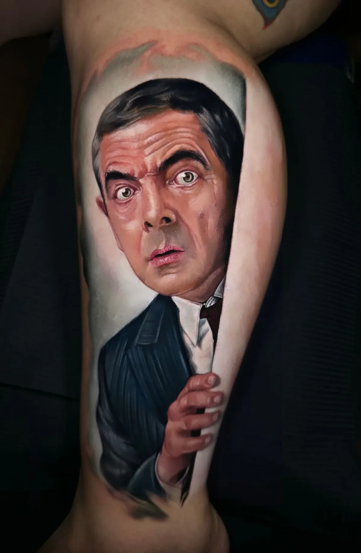A hyperrealistic tattoo depicting the face and upper torso of Mr. Bean with a wide-eyed bewildered expression, dark hair, pinstripe suit, and a flesh-toned limb curving into the frame creating a trompe-l'œil three-dimensional effect.