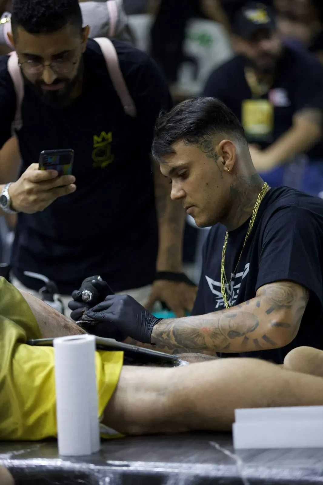 A tattoo artist with dark hair and gold jewellery, wearing black gloves, leans over a client's leg draped in yellow fabric and applies a tattoo; a man in glasses holds up a phone to record in the background.