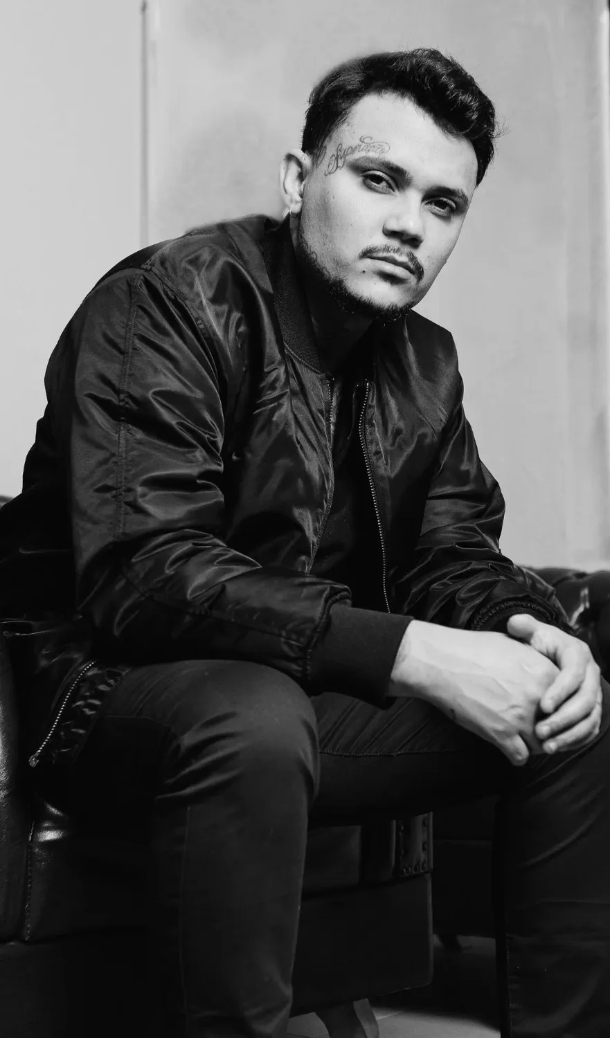 A black-and-white portrait of a young man with dark hair, a thin mustache and faint beard, seated on a dark leather couch wearing a zip-up bomber jacket, looking directly at the camera with a neutral expression; a small tattoo is visible above his left eyebrow.