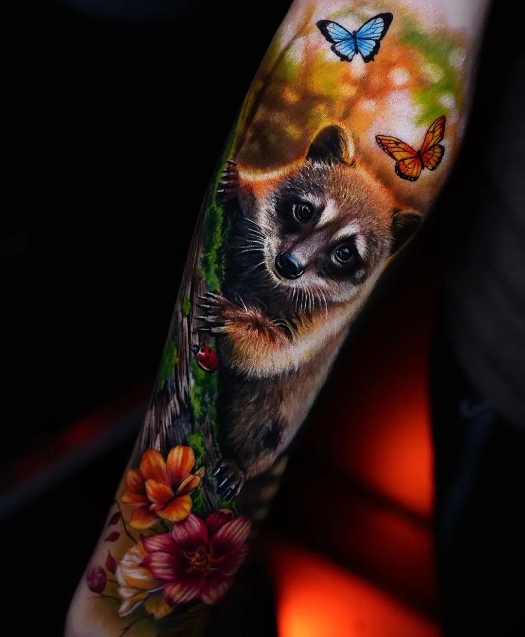 A photorealistic colour tattoo on a forearm showing a raccoon gripping a tree trunk; an orange monarch butterfly and a blue butterfly fly above, a red ladybug sits on the bark, and orange, red and yellow flowers bloom in the lower left corner.