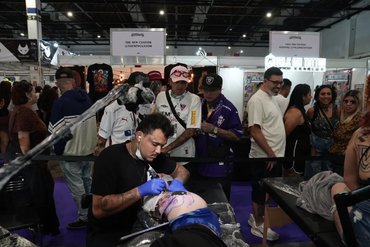 A tattoo artist wearing blue gloves and a black t-shirt works on a large intricate tattoo on a client's torso at a tattoo convention; a crowd of observers and branded booths fill the background.