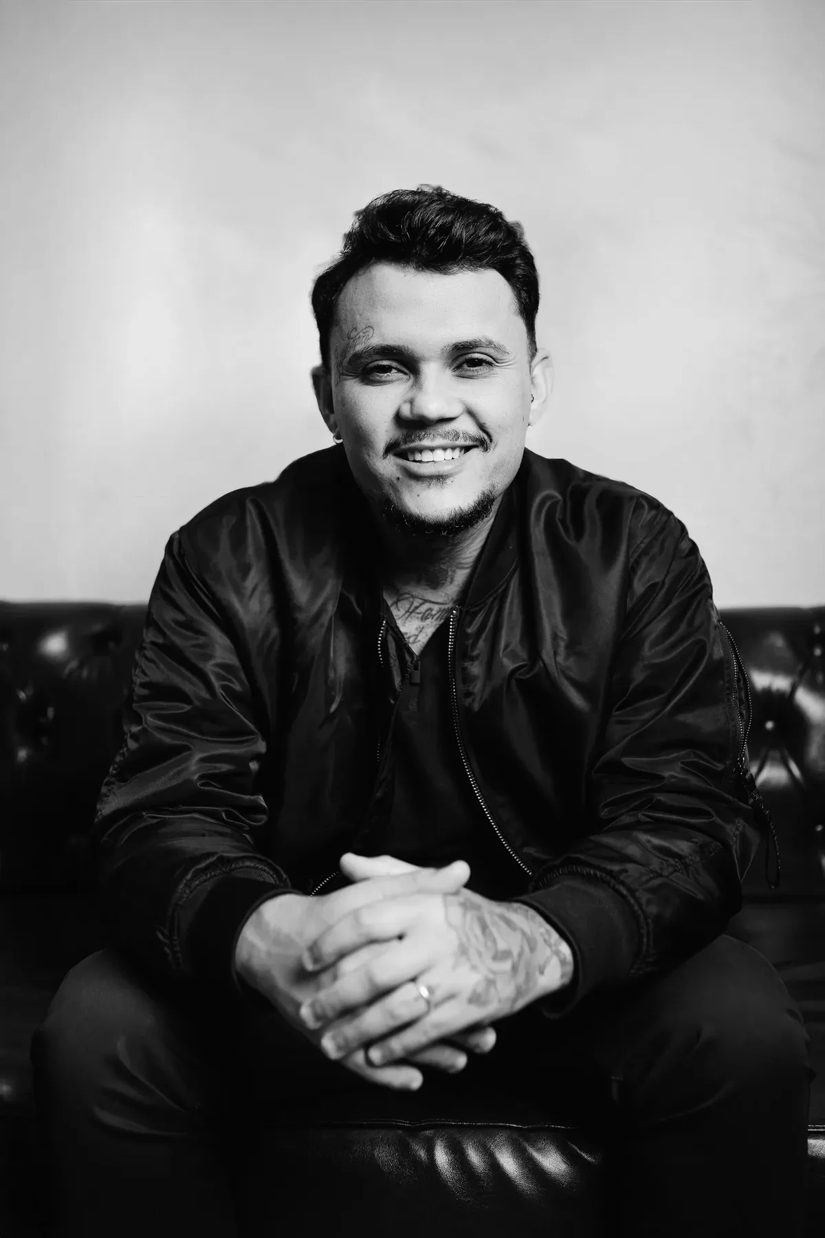 A black-and-white portrait of a young man with dark styled hair, a mustache and goatee, seated on a dark leather couch in a bomber jacket, smiling at the camera with his hands clasped in his lap; tattoos are visible on his hands and neck.