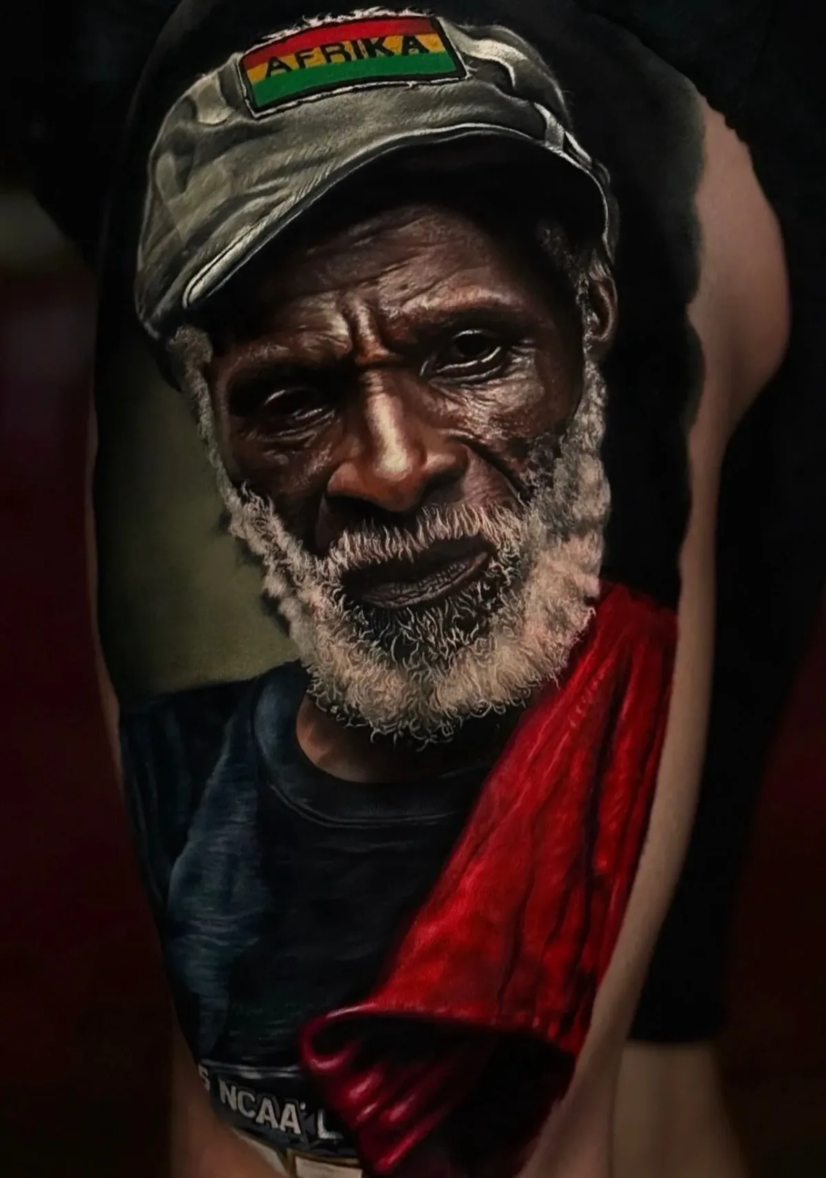 A hyperrealistic colour tattoo of an older Black man's face; he wears a grey cap with a rectangular Afrika patch in red, yellow and green, and a bright red cloth is draped over his shoulder, his grey beard rendered strand by strand.