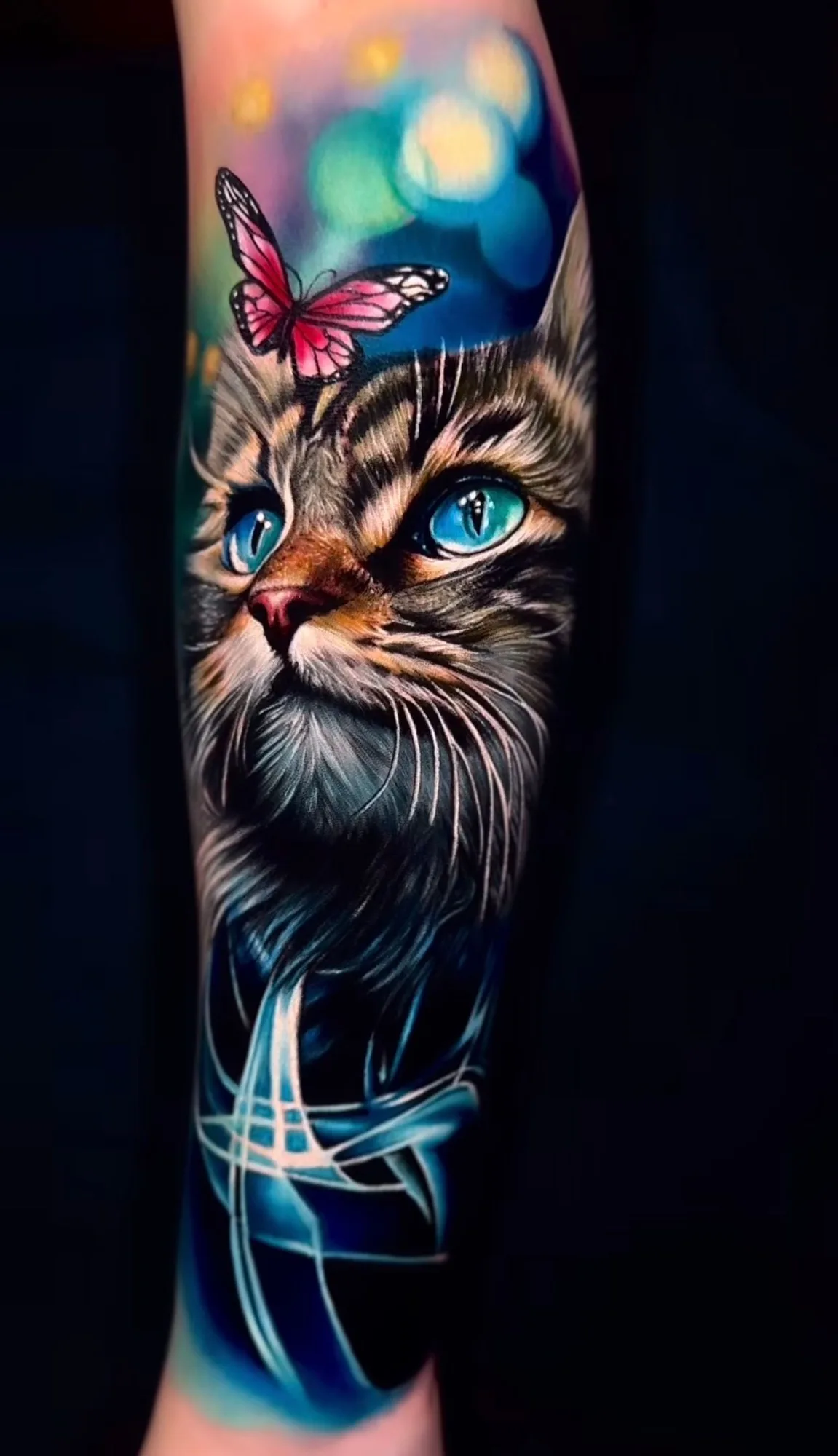 A hyperrealistic forearm tattoo of a cat's face with intense turquoise blue eyes and white whiskers, a small pink butterfly sitting on top of its head, and the lower portion of the tattoo transitioning into abstract swirling lines of electric blue and white against a dark background.