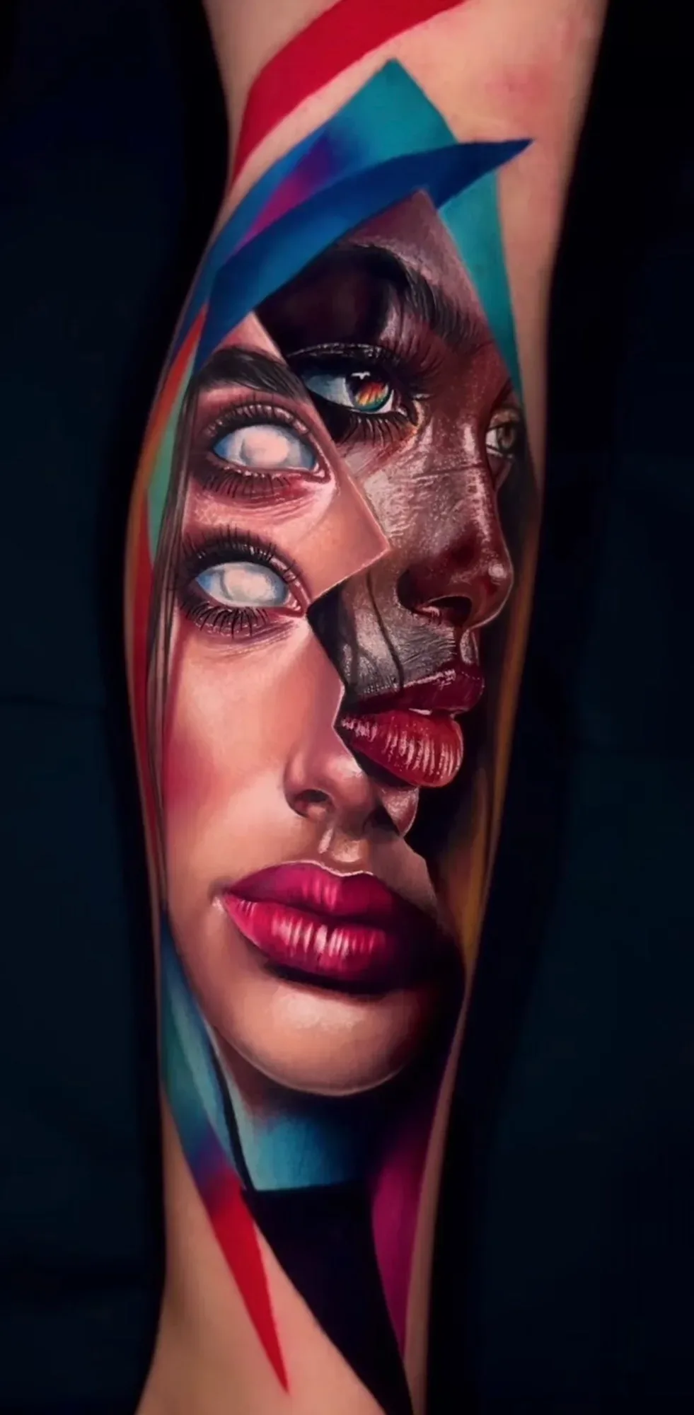 A hyperrealistic tattoo of a human face broken into angular fragments; multiple eyes, including ones with white irises, and full glossy red lips are visible across the shattered composition set against a black background with coloured geometric shapes.
