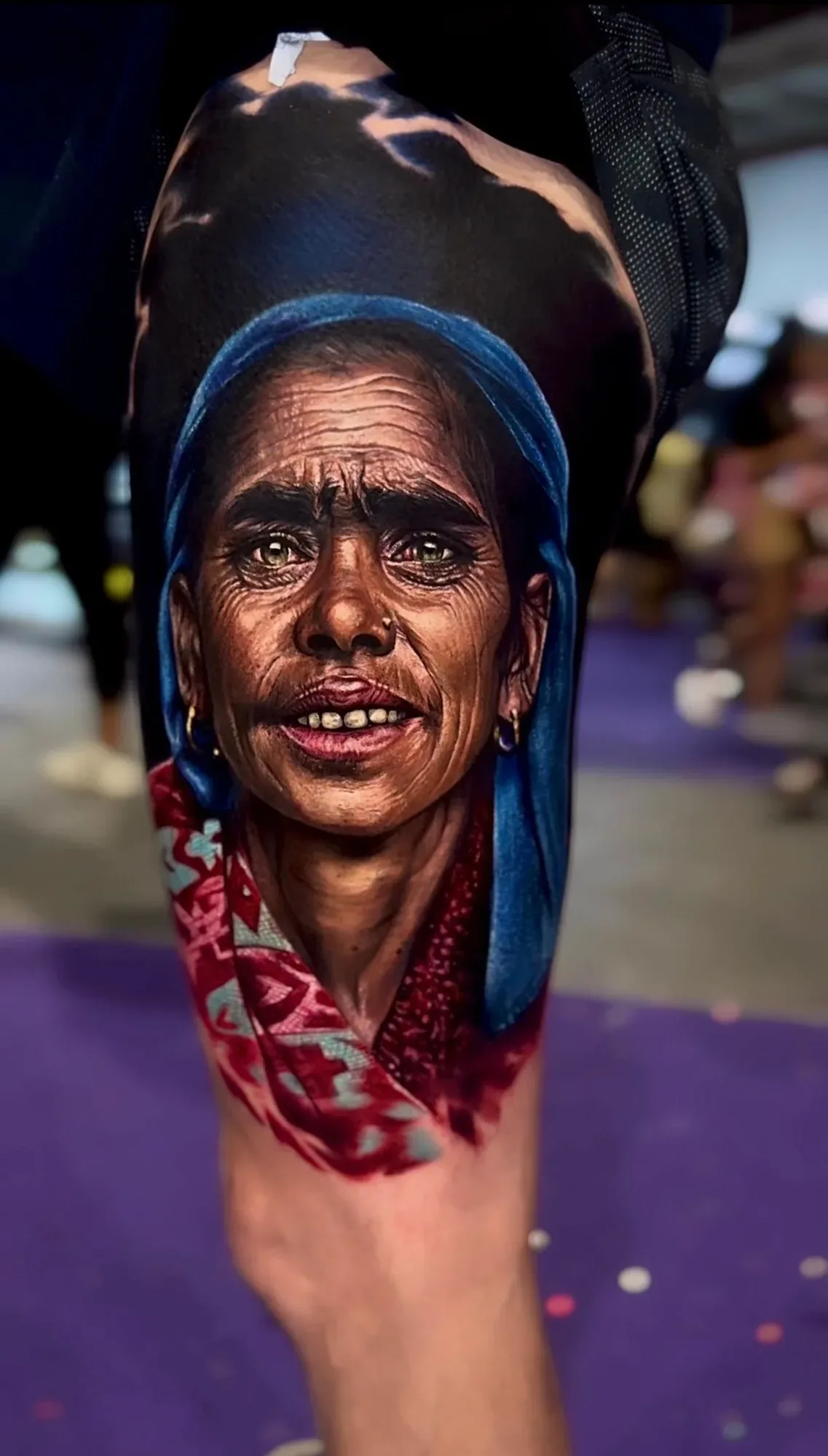 A hyperrealistic colour tattoo of an older woman's face; she wears a vivid blue headscarf, a red patterned fabric at her chest, small gold hoop earrings and a nose ring, her green eyes looking directly at the viewer.